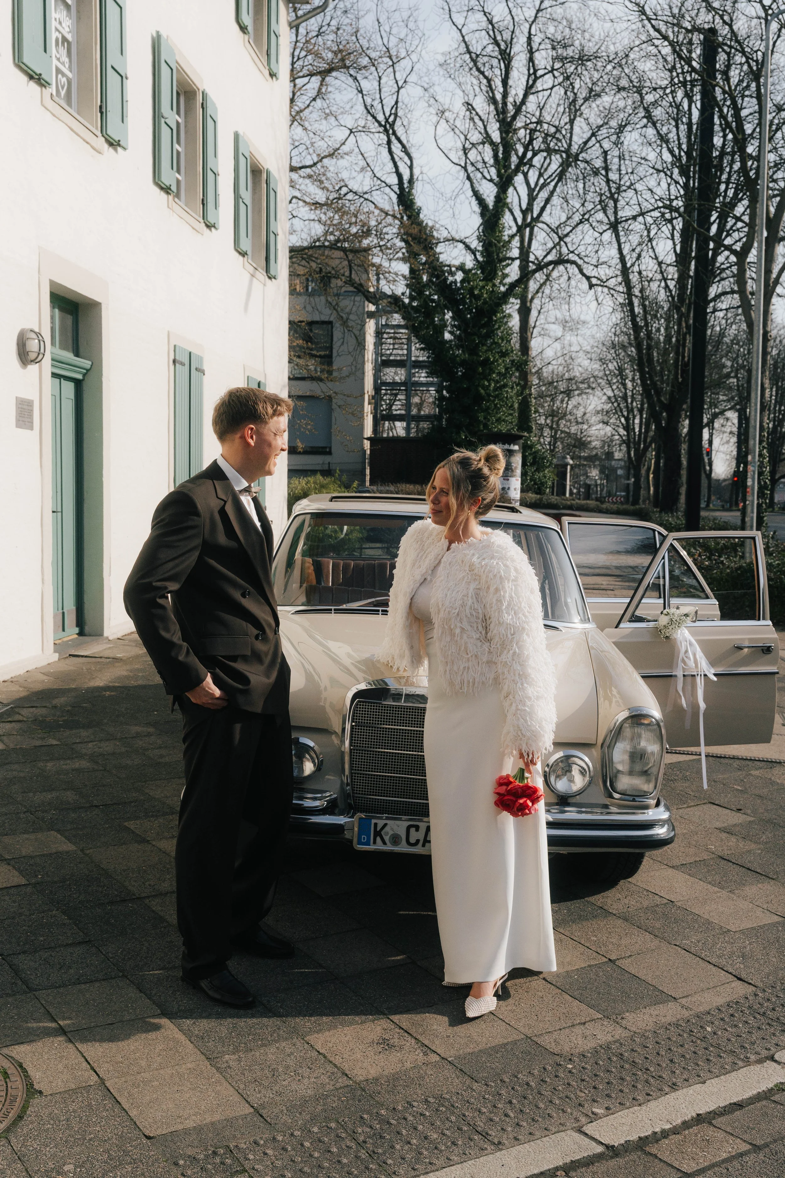 Ein Mann und eine Frau stehen vor einem weißen Auto, wahrscheinlich an einem Hochzeitstag. Die Frau trägt ein langes weißes Kleid und hält rote Rosen, während der Mann einen schwarzen Anzug trägt.