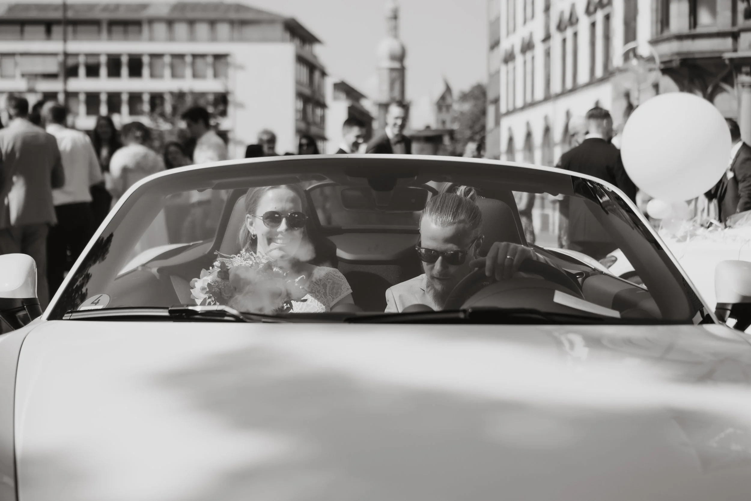 Braut und Bräutigam in einem klassischen Cabrio bei einer Hochzeitsfeier, umgeben von Gästen in einer Stadtstraße. Das Bild ist in Schwarz-Weiß aufgenommen.