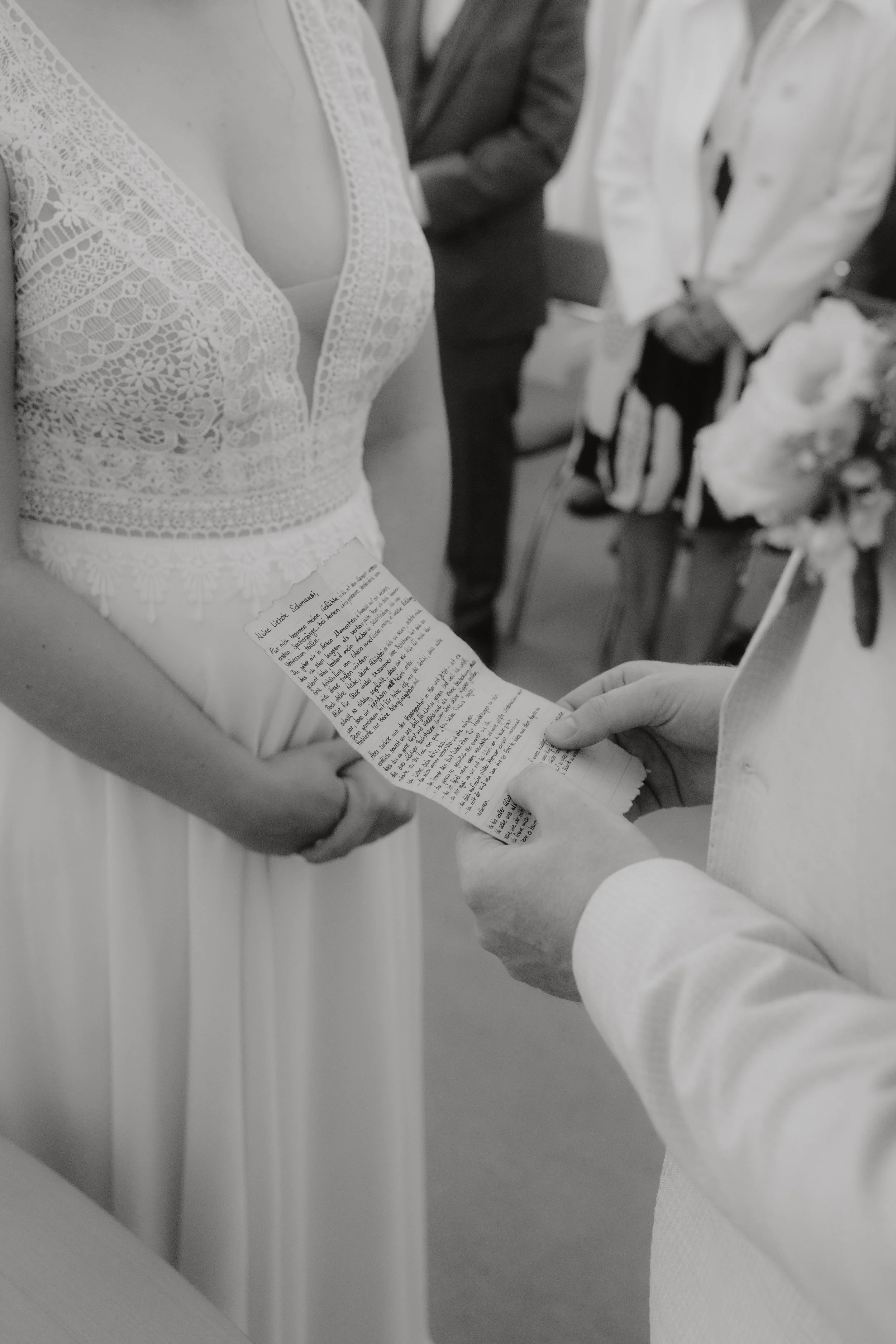 Ein Brautpaar bei einer Hochzeit, die Braut trägt ein weißes Kleid mit Spitze und liest ein gelbes Papier, vermutlich eine Liebeserklärung oder einen Eheversprechen, während der Bräutigam im weißen Anzug steht.