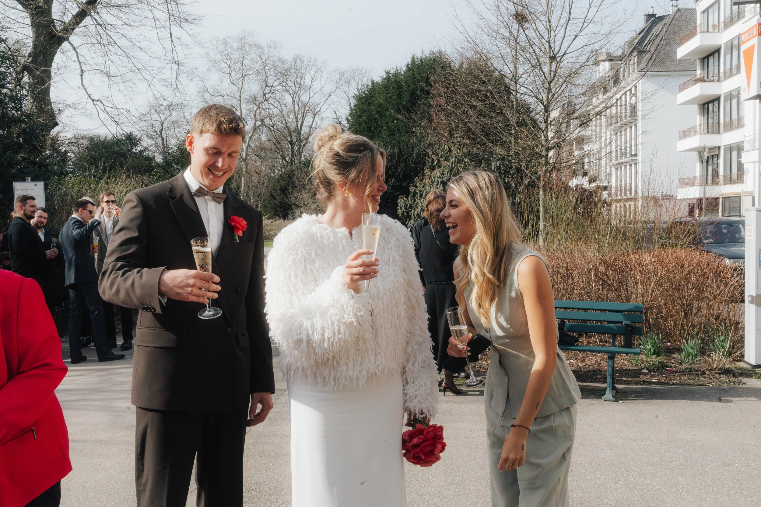 Menschen bei einer Hochzeit im Freien, unterhalten sich und trinken Sekt, im Hintergrund einige Gäste, Bäume und moderne Gebäude.