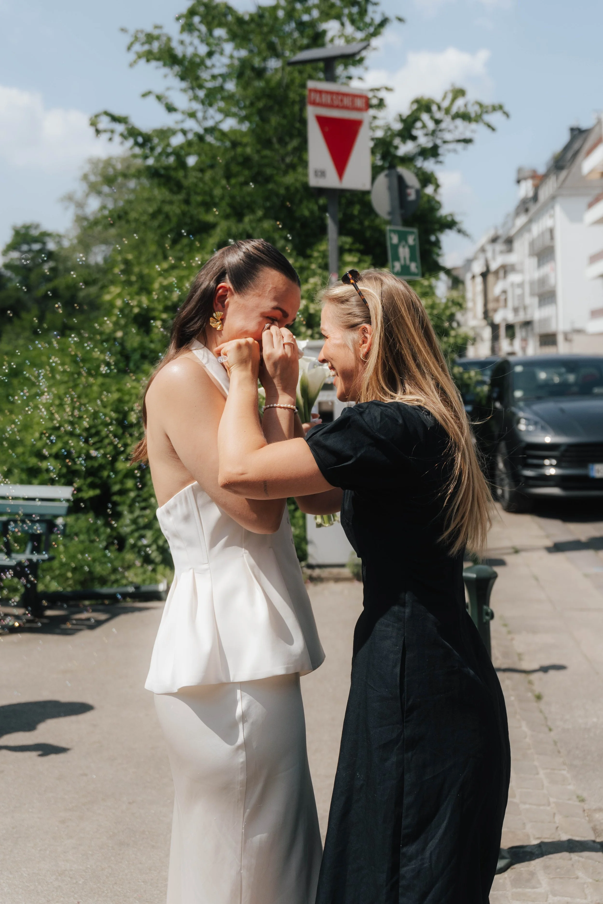 Zwei Frauen, eine in einem weißen Kleid, die andere in schwarzen Kleid, lachen und umarmen sich auf der Straße bei sonnigem Wetter, im Hintergrund Bäume, Gebäude und Autos, während sie sich freuen und glücklich sind.