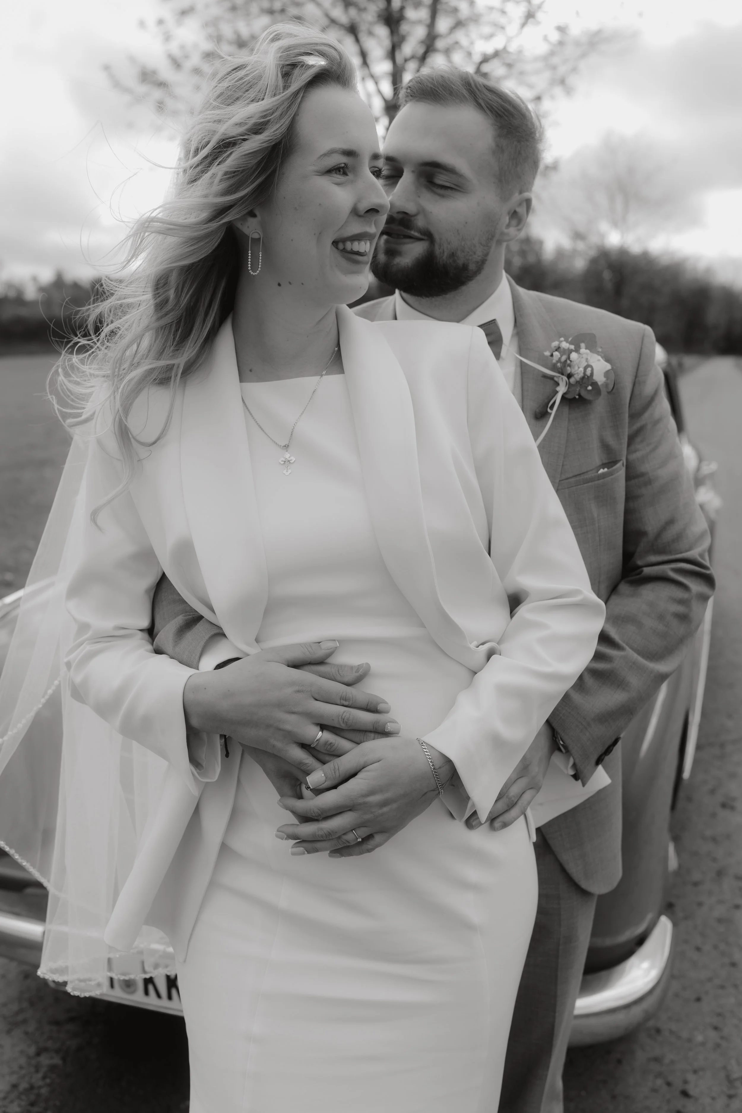 Ein Brautpaar bei Hochzeit, in Schwarz-Weiß, lächelnd, mit Liebe verbunden, an einem Tag im Freien, mit einem Auto im Hintergrund.