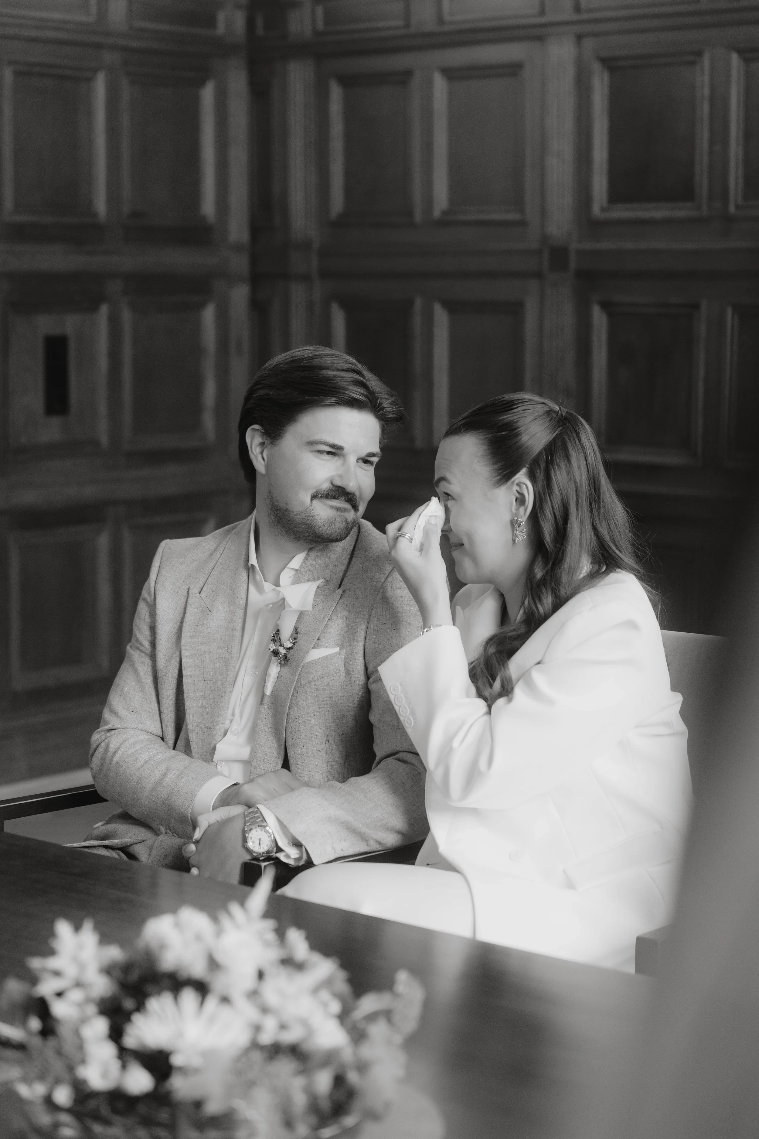 Ein Mann und eine Frau, die bei einer Hochzeit weinen, sitzen an einem Tisch in einem Raum mit dunklen Holzwänden.