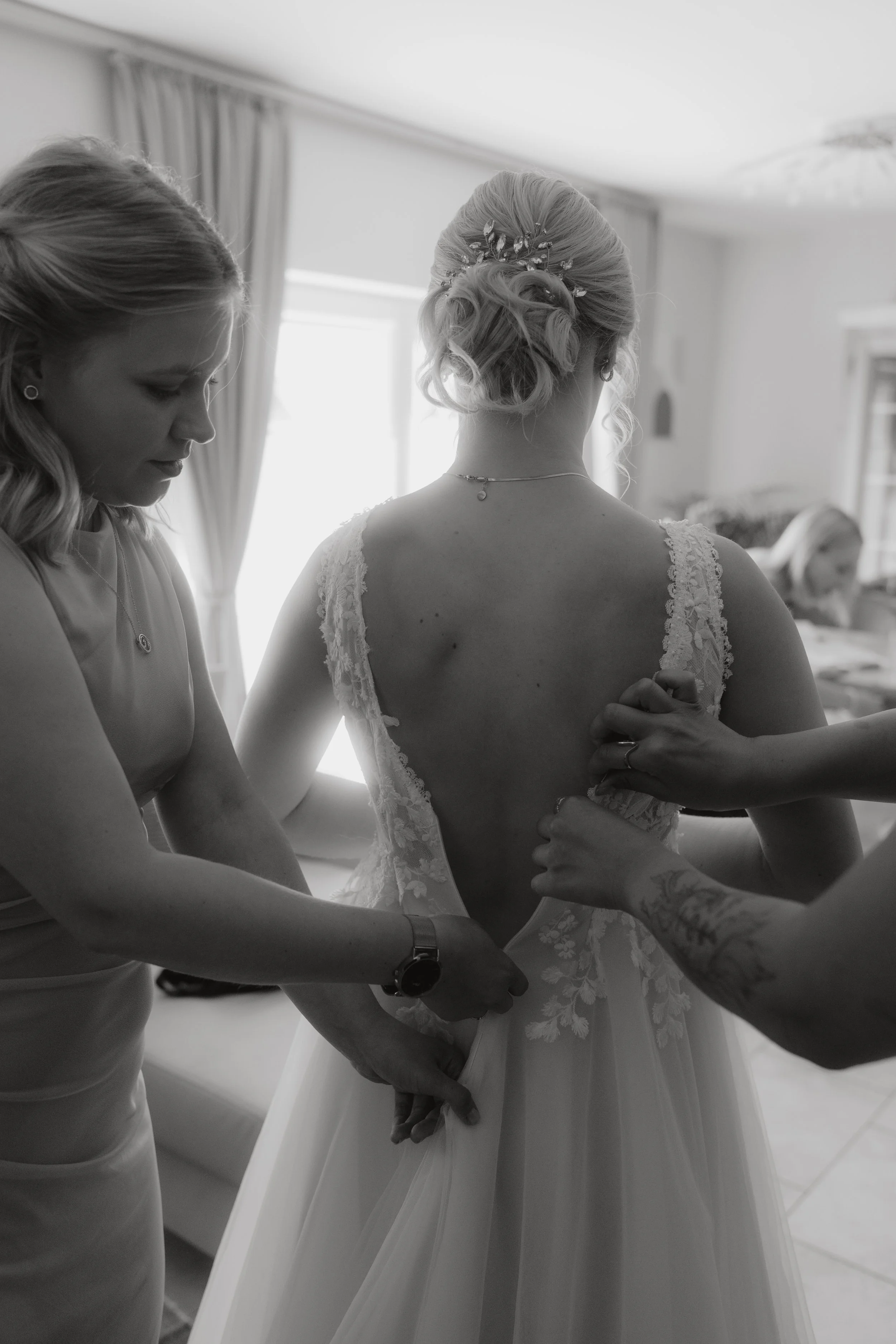 Braut wird in einem Zimmer für die Hochzeit vorbereitet, während Helferinnen beim Anziehen ihres Brautkleides assistieren.