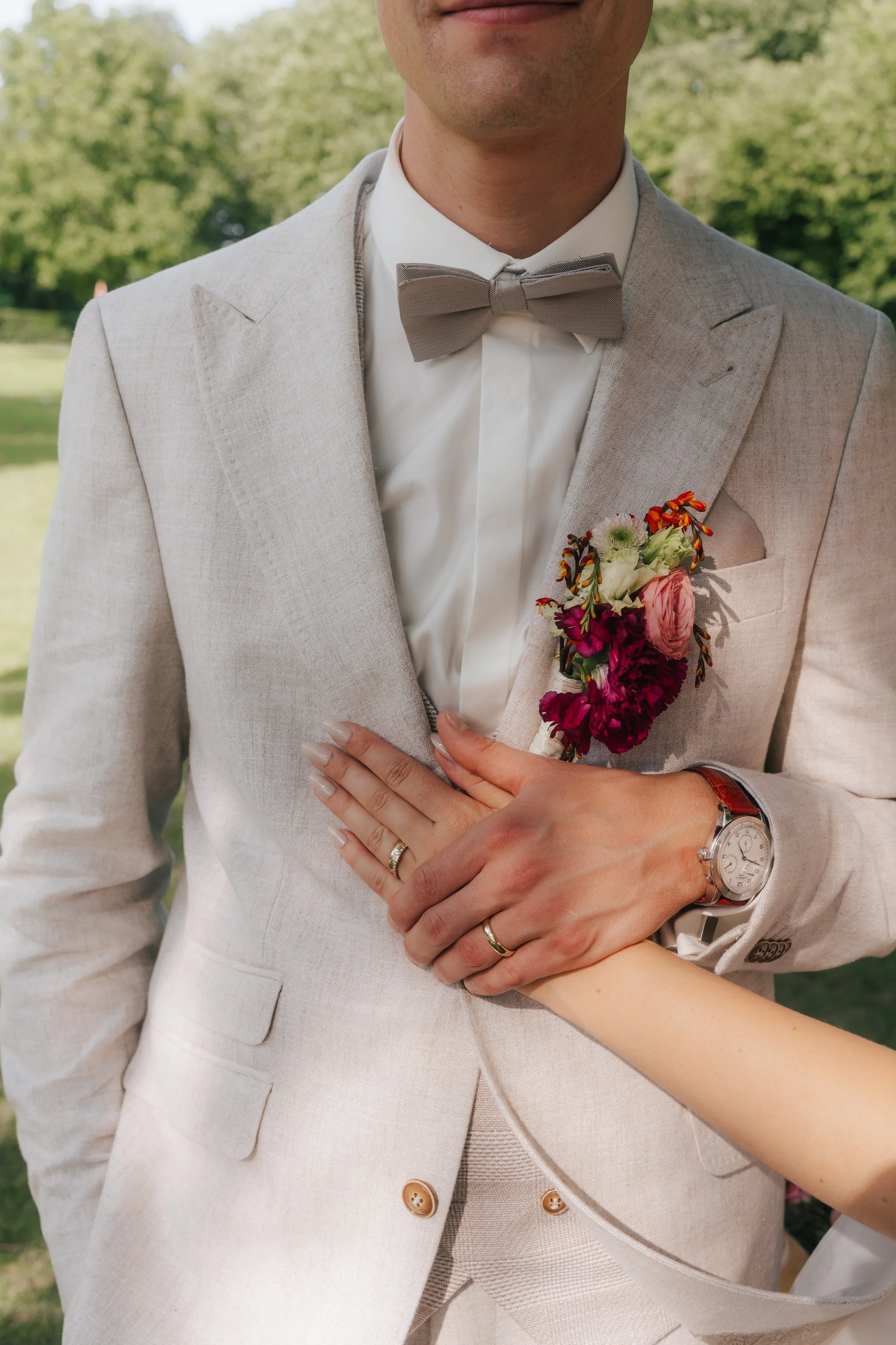 Nahaufnahme eines Mannes in einem hellen Anzug mit Fliege, ein Blumenstrauß im Revers und eine Uhr am Handgelenk, bei einer Hochzeit im Freien.