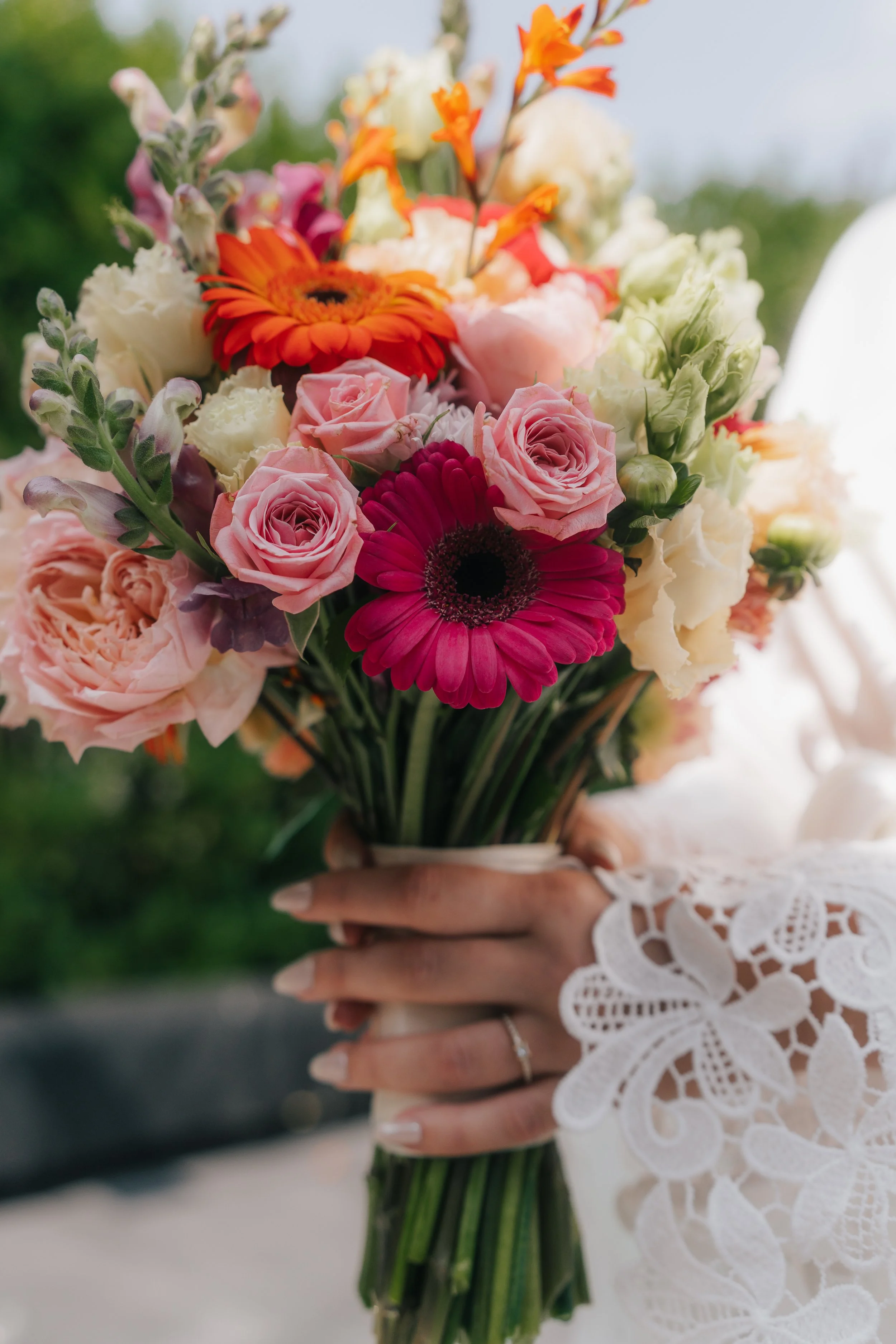 Eine Hand hält einen bunten Blumenstrauß mit rosa Rosen, orange und dunkelorangen Gerbera, weißen und lilafarbenen Blumen, vor einem grünen, unscharfen Hintergrund.