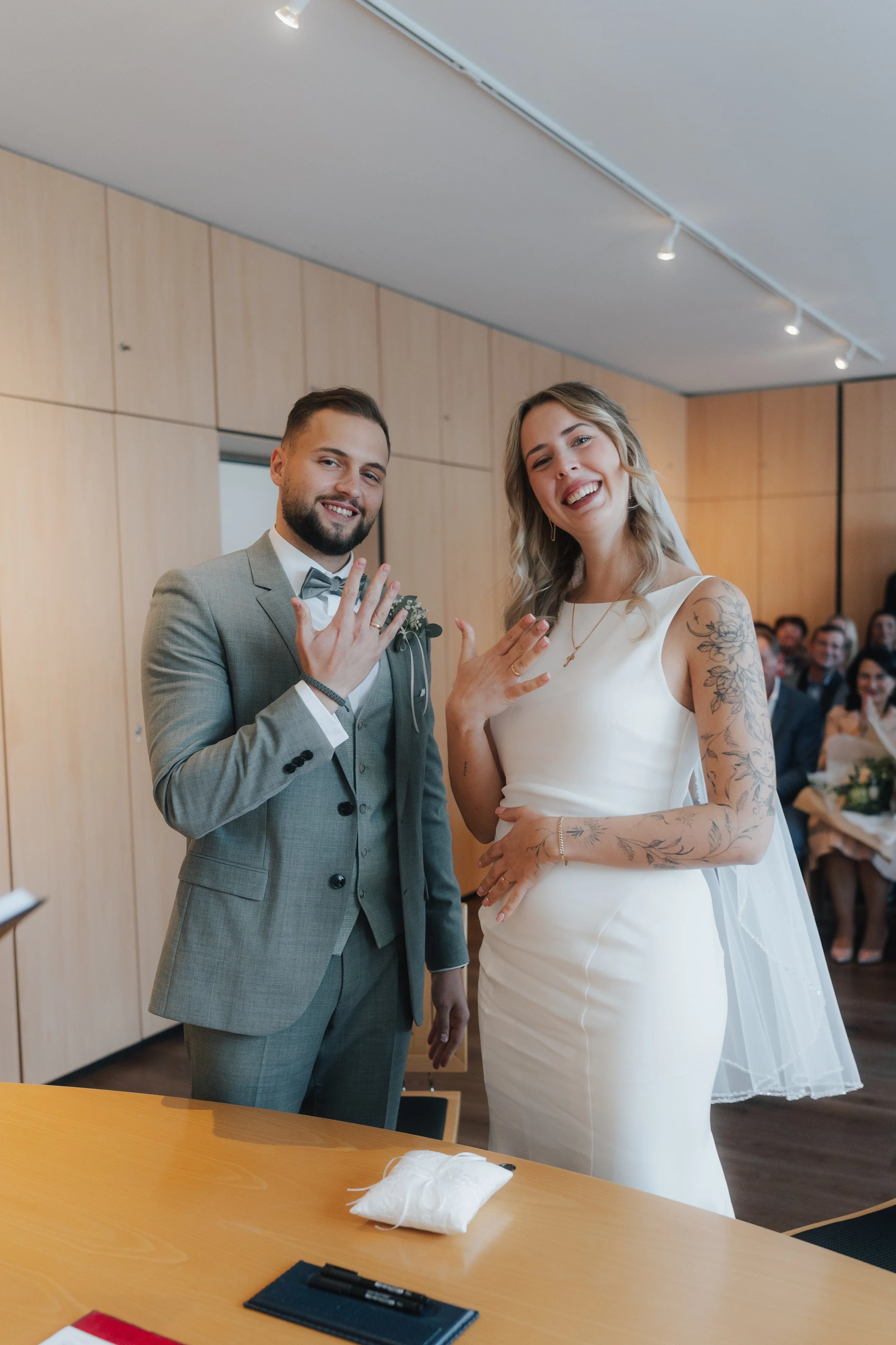 Ein Brautpaar zeigt ihre Eheringe bei einer Hochzeit. Die Frau trägt ein weißes Brautkleid mit Tattoos am Arm, der Mann trägt einen grauen Anzug.
