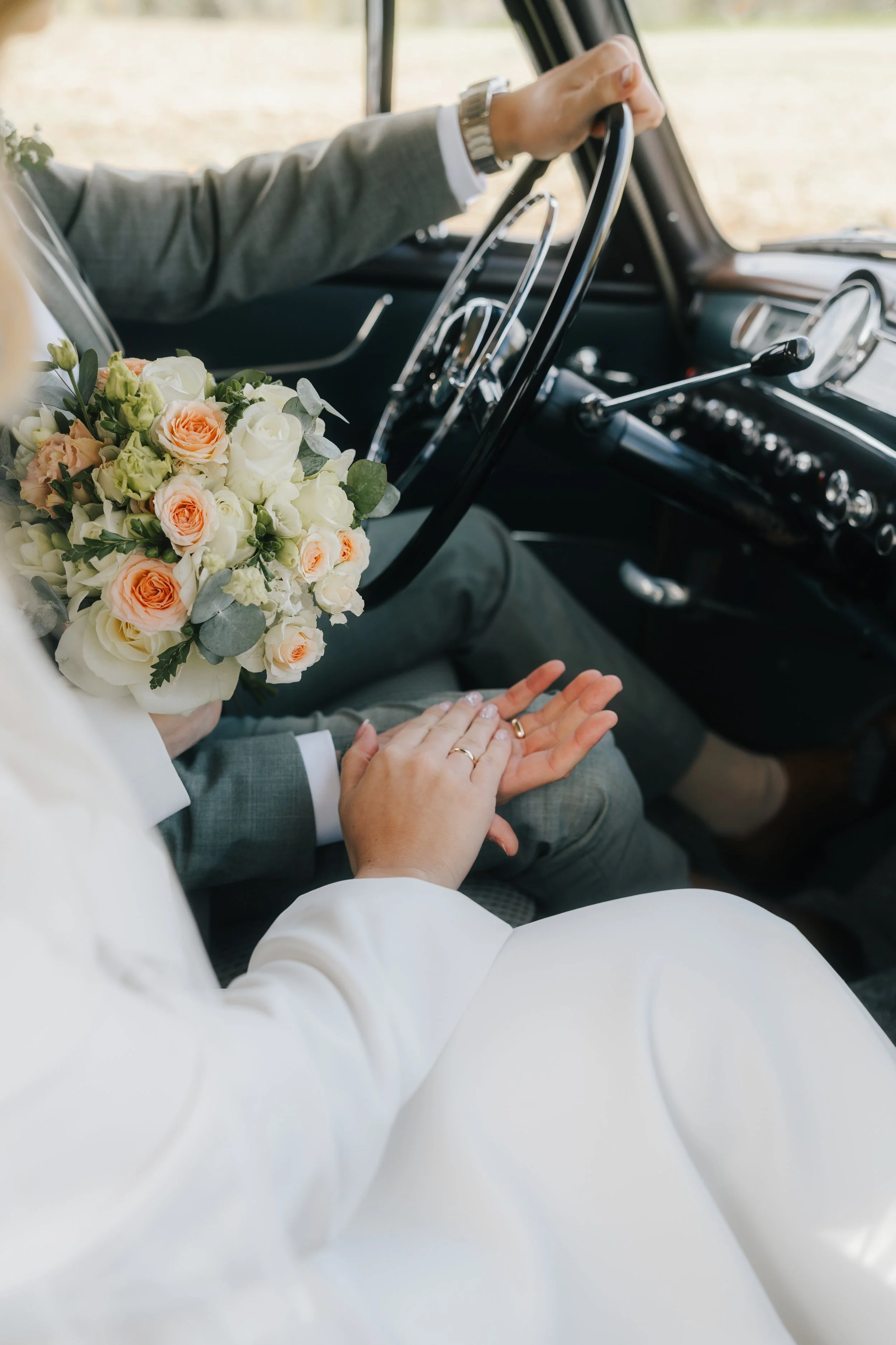 Ein Brautpaar hält sich die Hände im Inneren eines Autos. Die Braut trägt ein weißes Kleid und hält einen Blumenstrauß, während der Bräutigam Anzug trägt.
