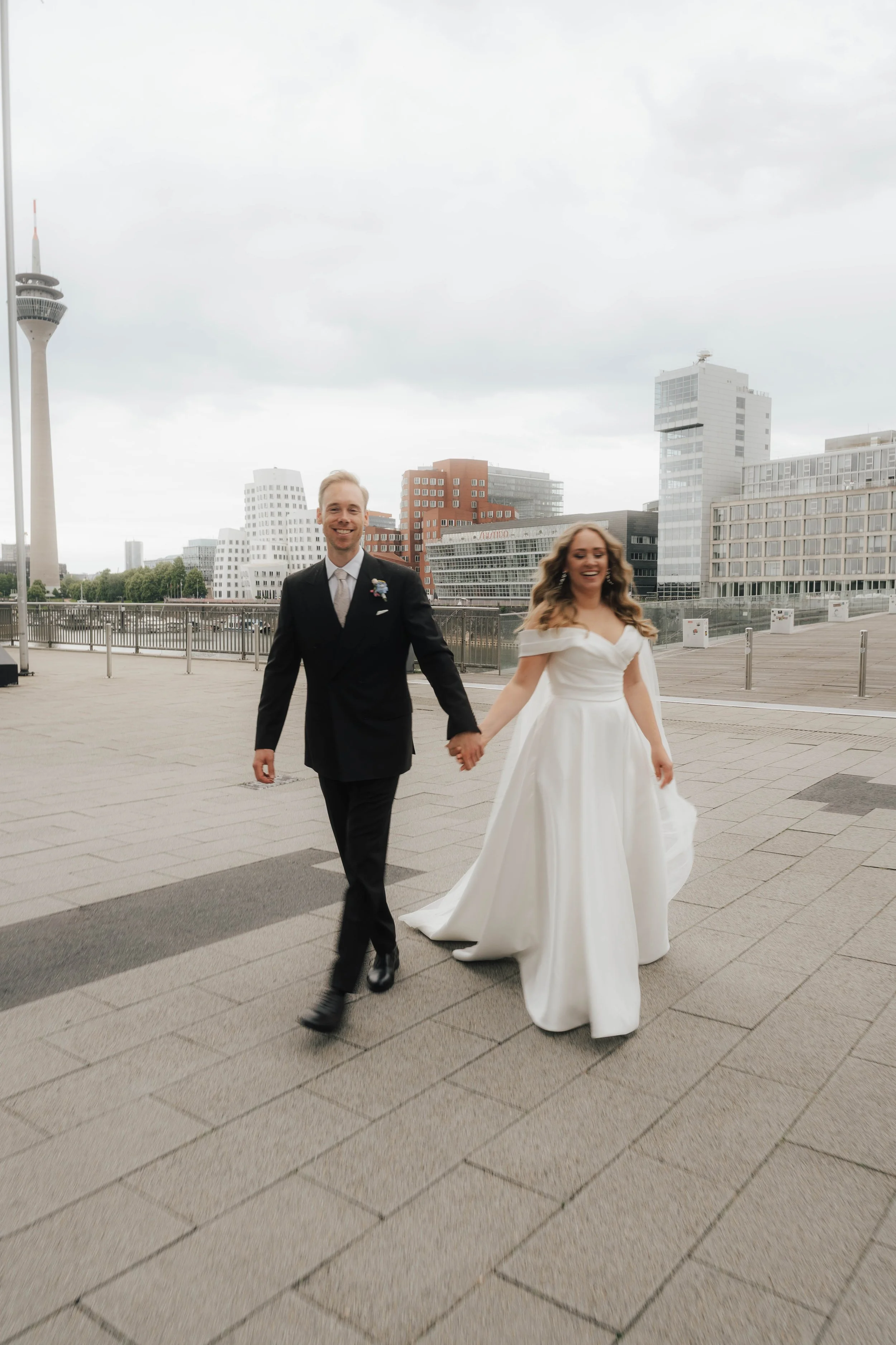 Ein glückliches Paar bei ihrer Hochzeit spaziert in einer modernen Stadt mit Hochhäusern und einem Fernsehturm im Hintergrund, während sie die Hand halten.