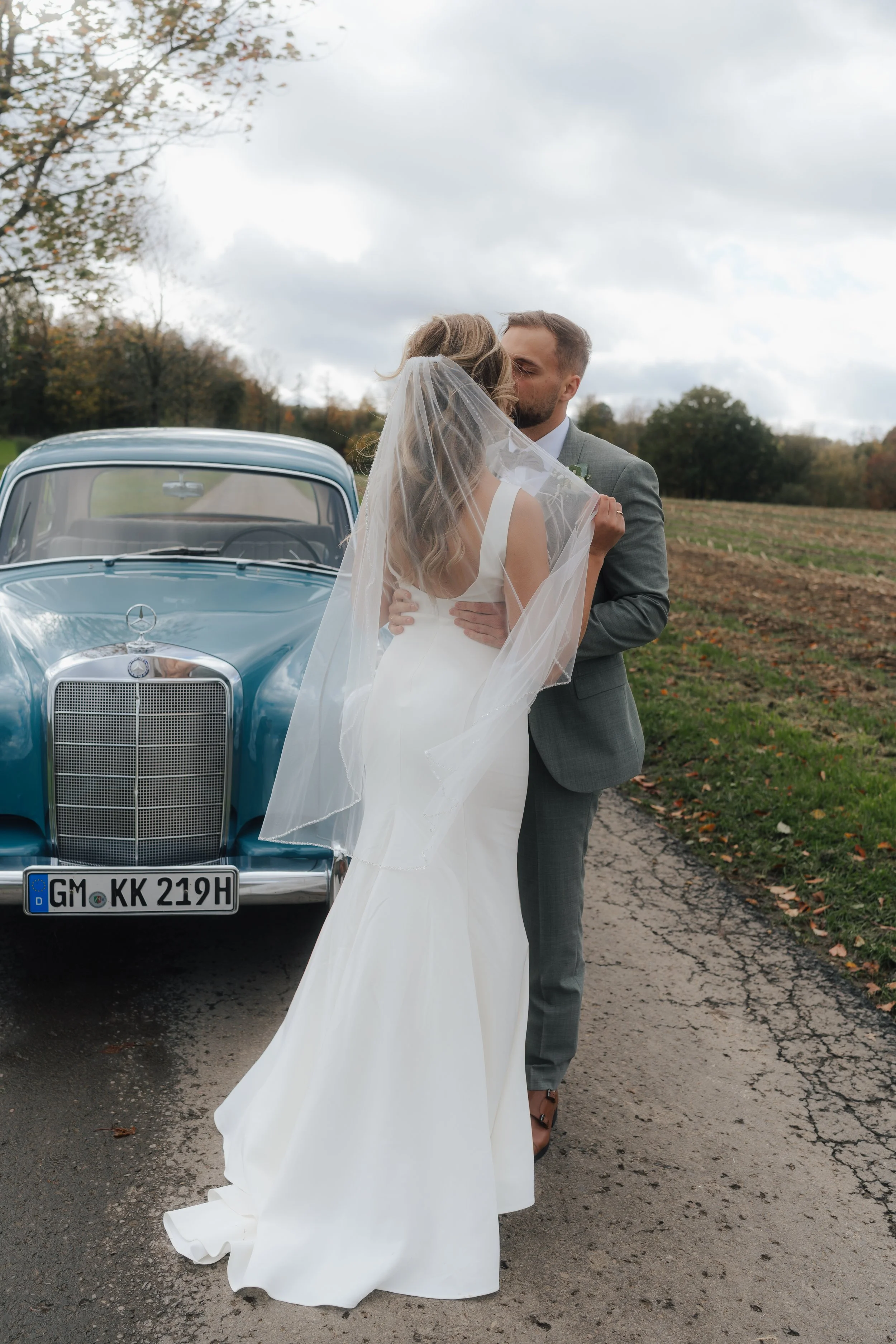 Ein Brautpaar küsst sich vor einem klassischen blauen Mercedes-Benz Auto auf einer Landstraße bei bewölktem Himmel.