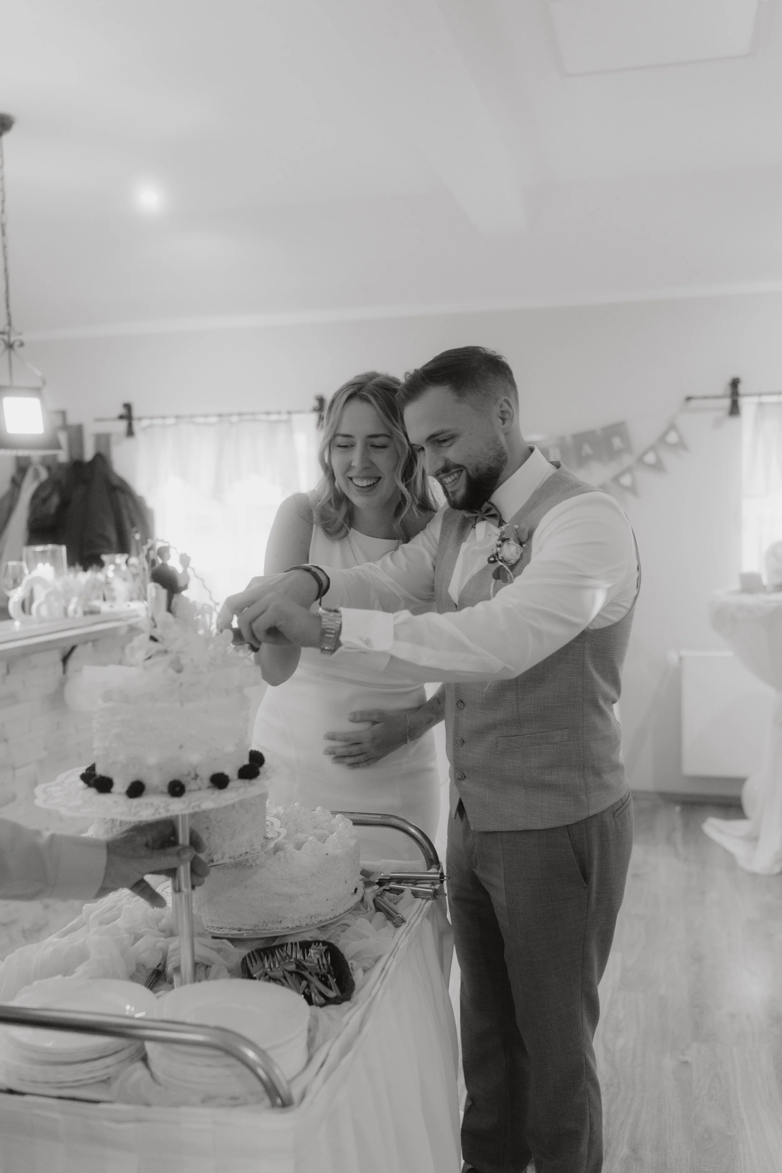 Ein Brautpaar schneidet gemeinsam eine Hochzeitstorte bei einer Feier. Die Frau trägt ein weißes Kleid, der Mann einen Anzug mit Fliege. Es gibt festliche Dekorationen im Raum.