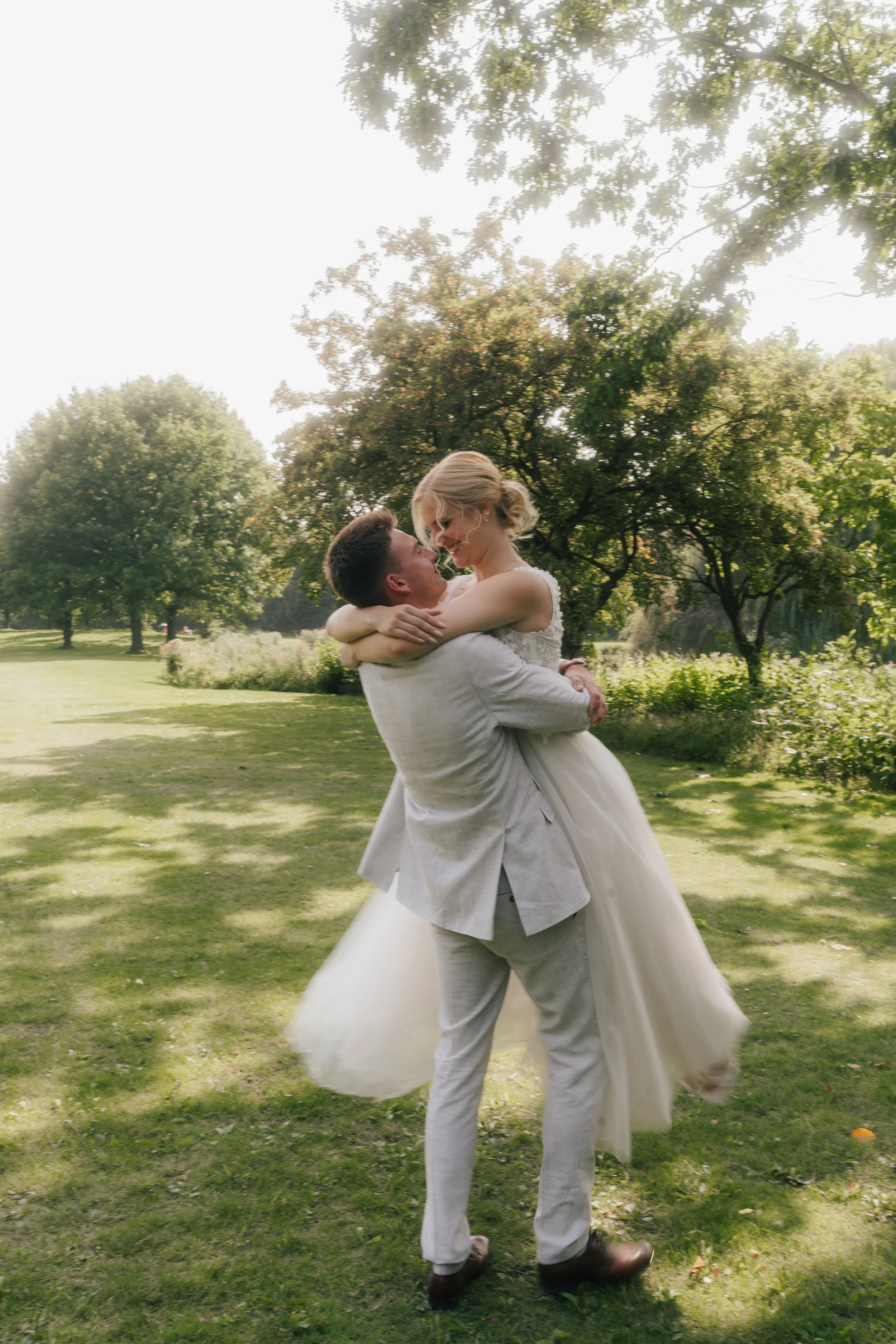 Ein Paar, das auf einer Wiese im Park tanzt, bei einem Hochzeitsfoto, lächelnd in die Augen schauend, umgeben von Bäumen und Sonnenschein.