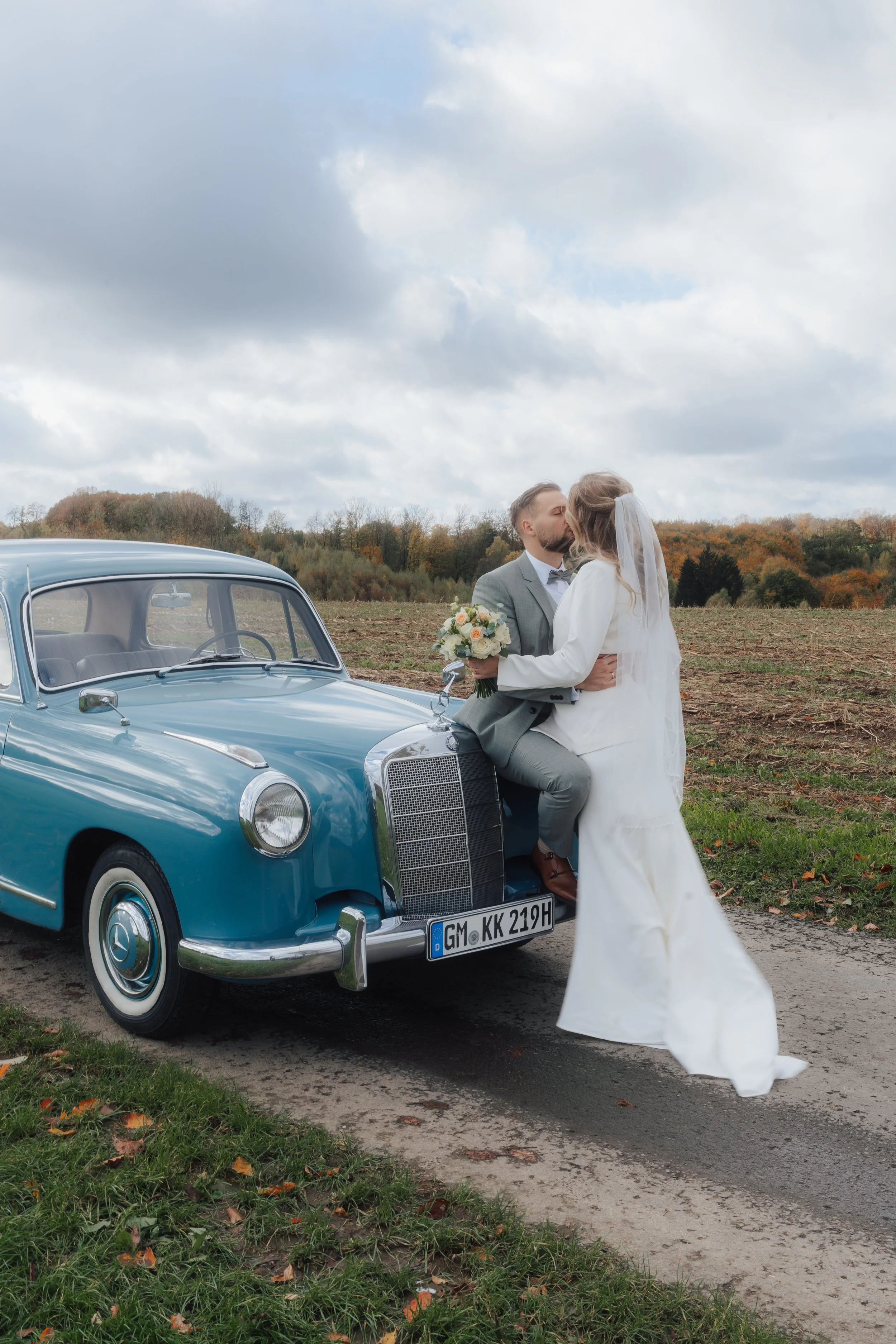 Ein Brautpaar küsst sich vor einem Vintage-Mercedes auf einem Feld bei bewölktem Himmel.