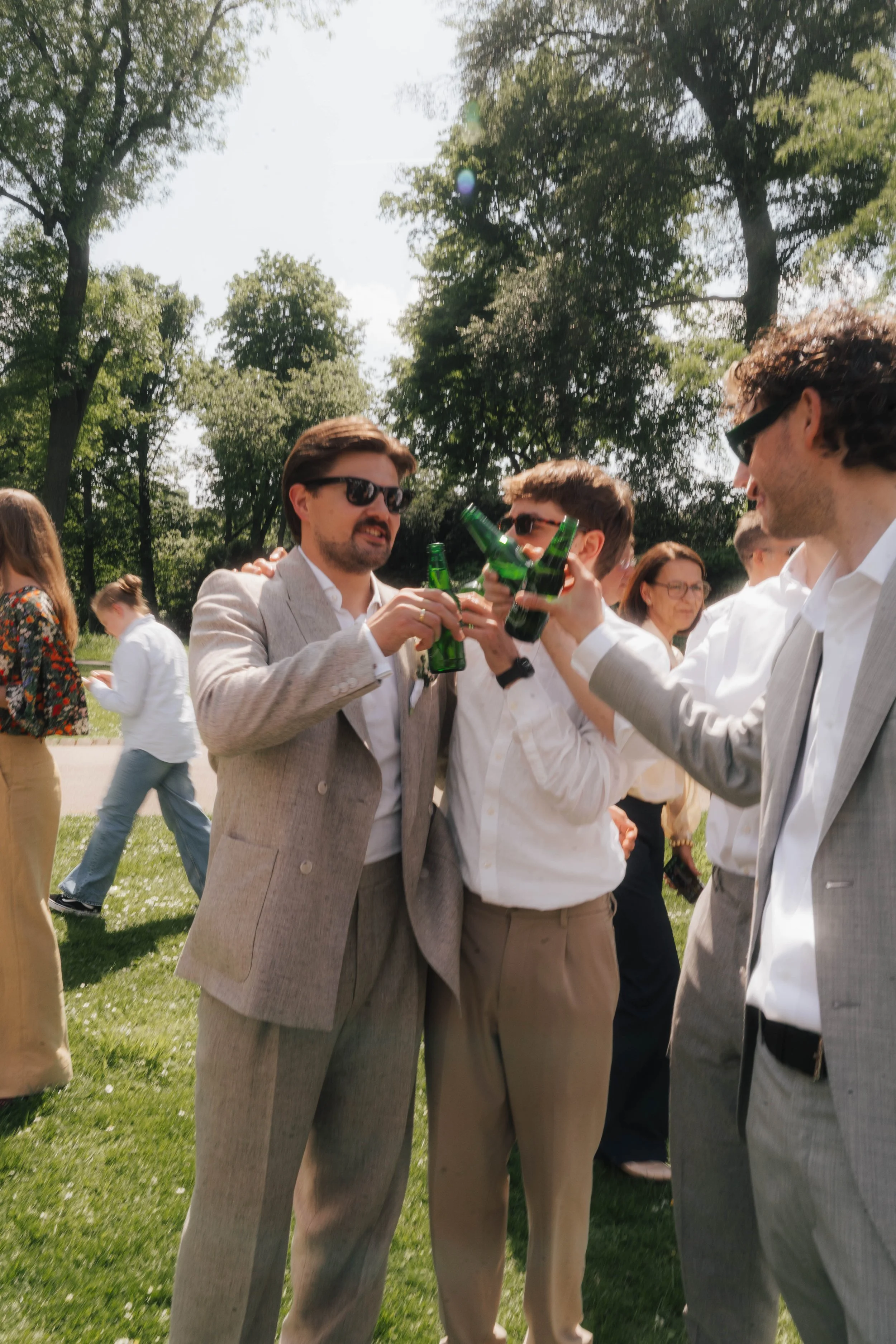 Mehrere Männer, die in Anzügen und Sonnenbrillen an einem sonnigen Tag im Park fröhlich mit Bierflaschen anstoßen.