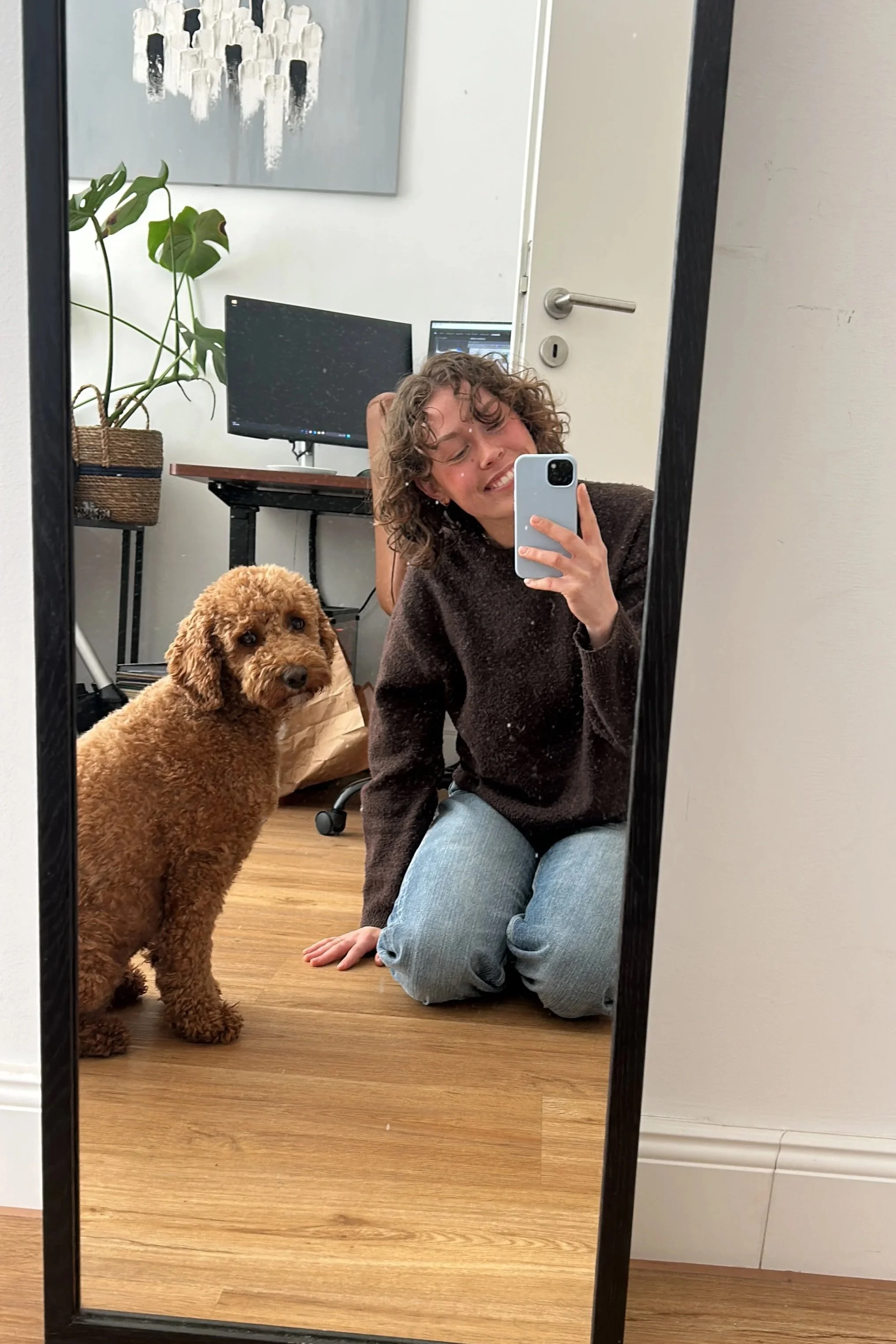 Spiegelbild einer Person mit lockigem Haar, die auf dem Boden kniet, neben einem goldenbraunen Hund, in einem Raum mit Computerbildschirm, Wandkunst und Pflanzen, fotografiert mit einem Smartphone.
