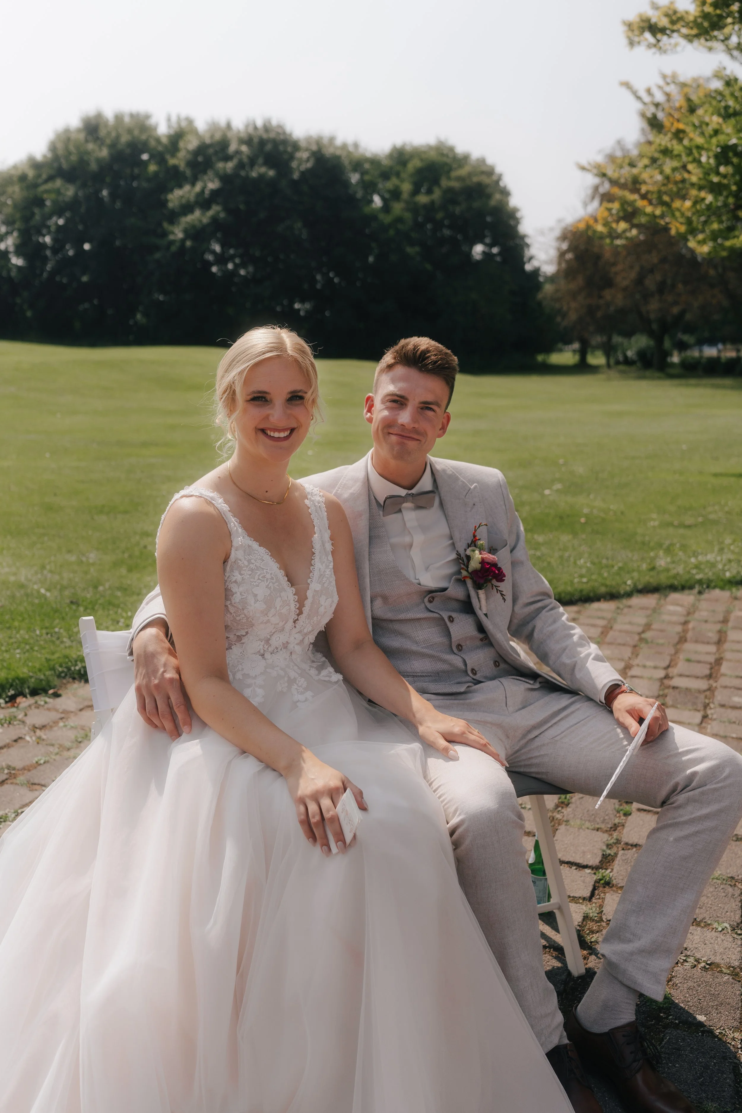 Ein Brautpaar sitzt auf einer Bank im Freien an einem sonnigen Tag, die Braut trägt ein weißes Hochzeitskleid und der Bräutigam ein graues Anzug mit Krawatte.