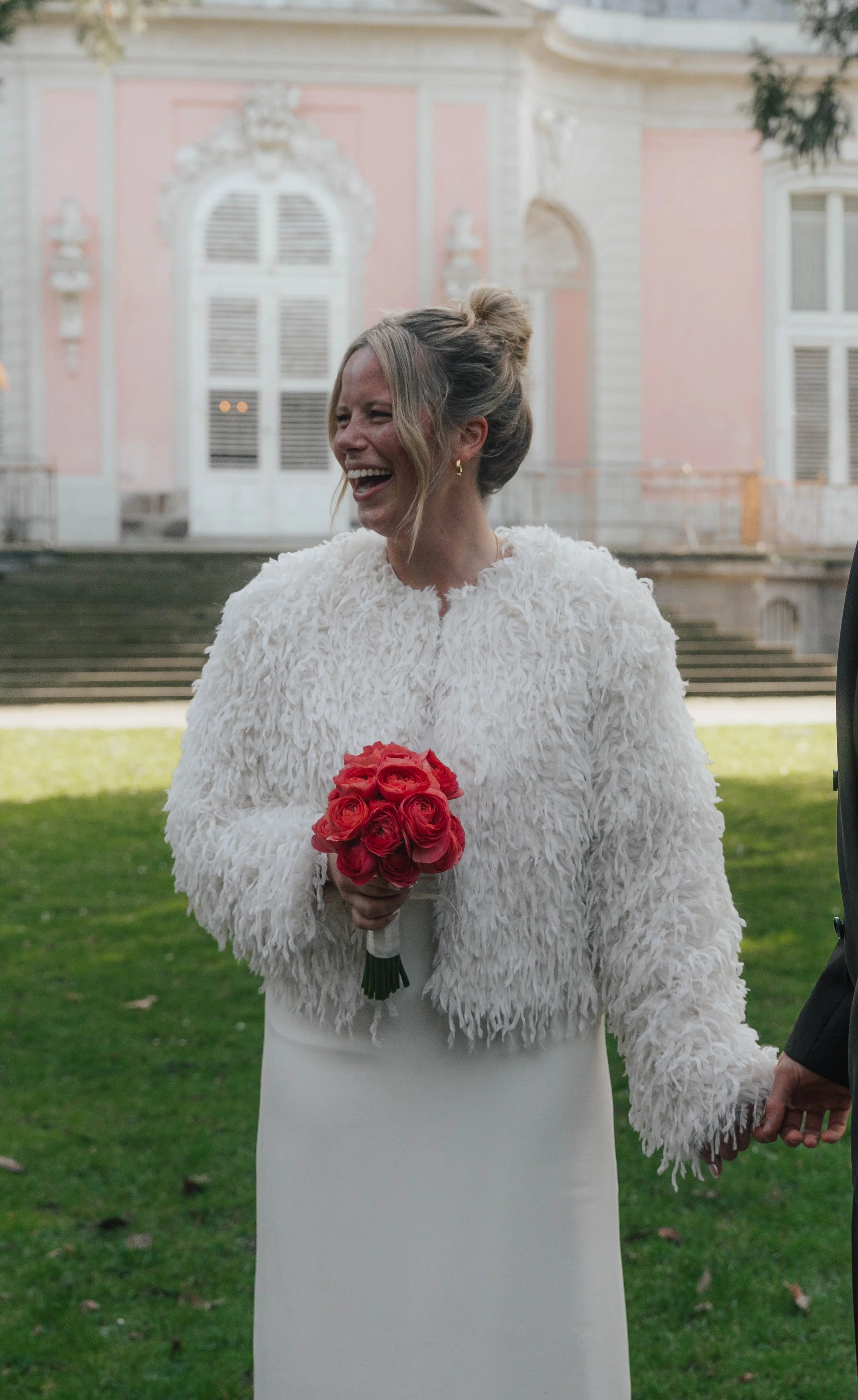 Junge Frau in einem weißen Kleid und einer weißen Felljacke hält einen roten Rosenstrauß in den Händen und lacht draußen vor einem historischen Gebäude.