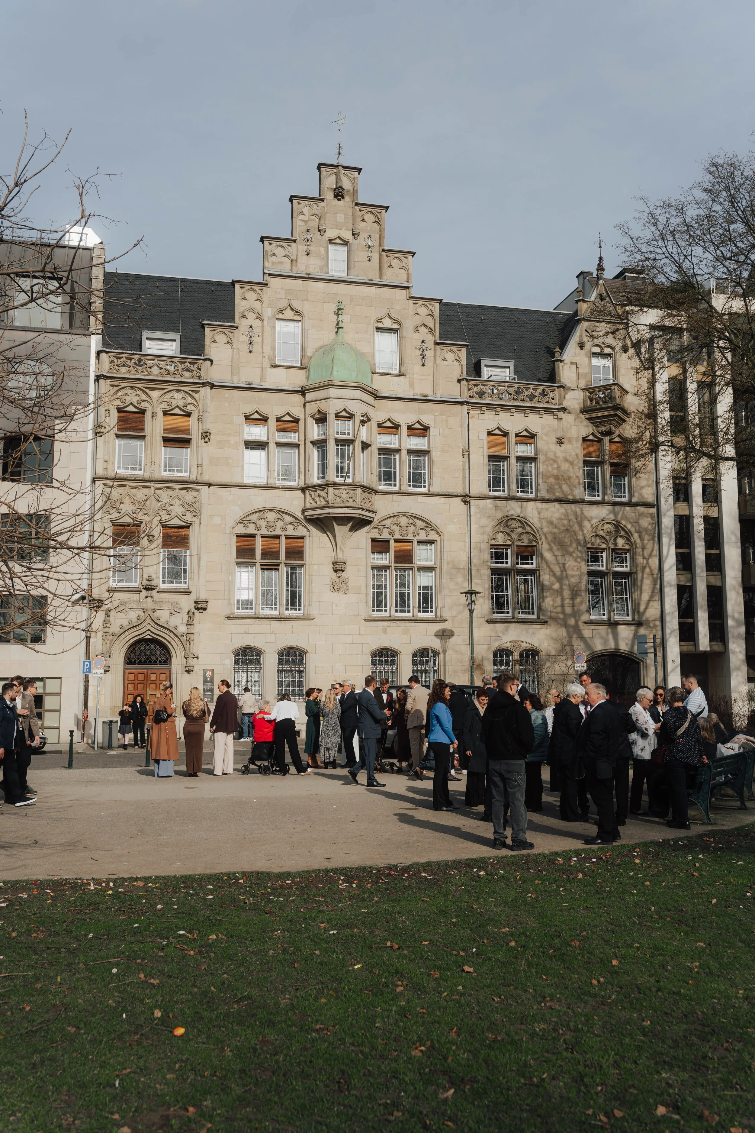 Menschen versammelt sich im Freien vor einem historischen Gebäude mit gotischer Architektur, auf einem Platz mit Gras und Bäumen.