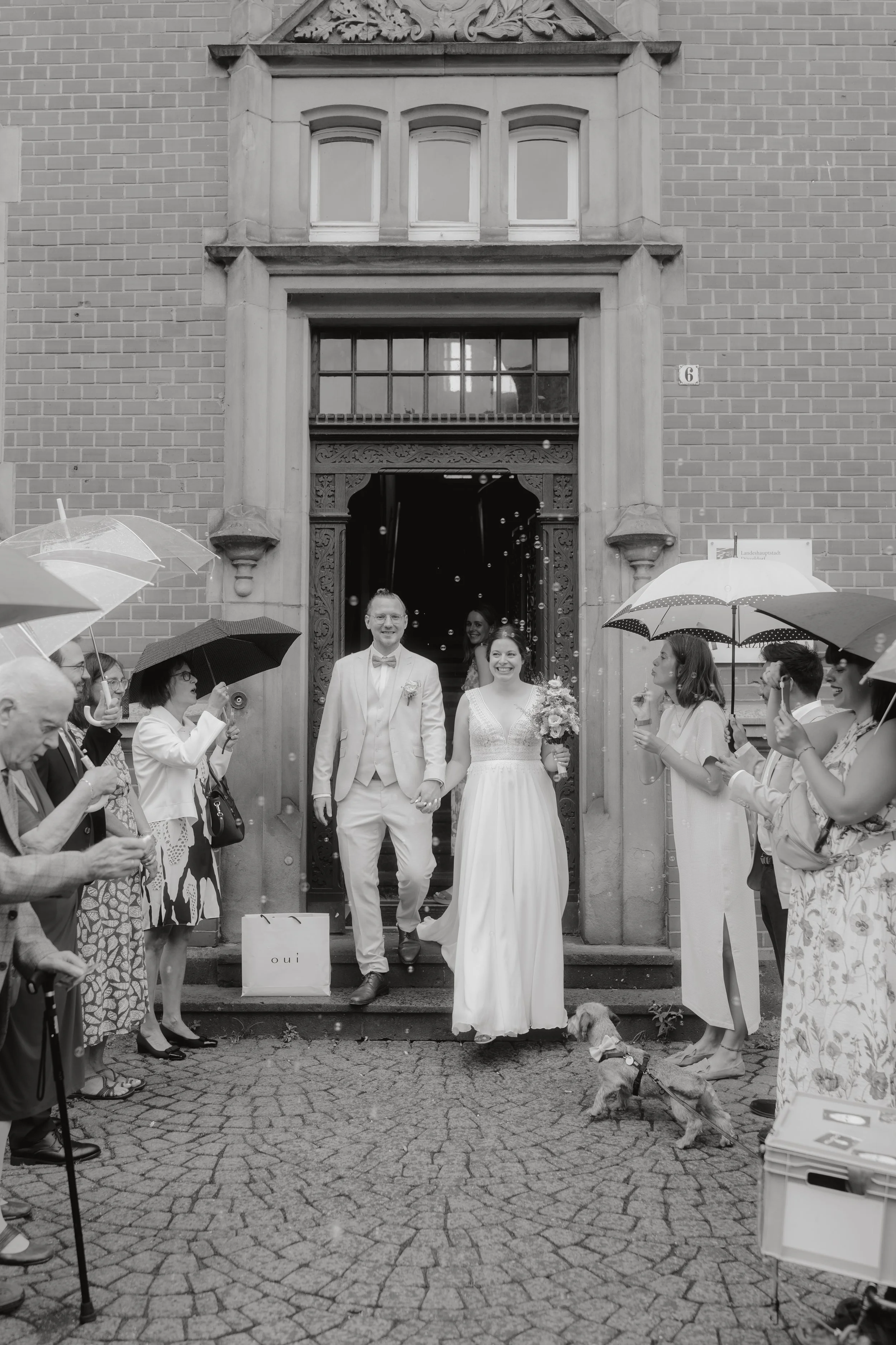 Braut und Bräutigam kommen strahlend aus einer Kirche, umgeben von Gästen mit Regenschirmen, die ihnen Glückwünsche bereiten, während sie Blumen streuen.