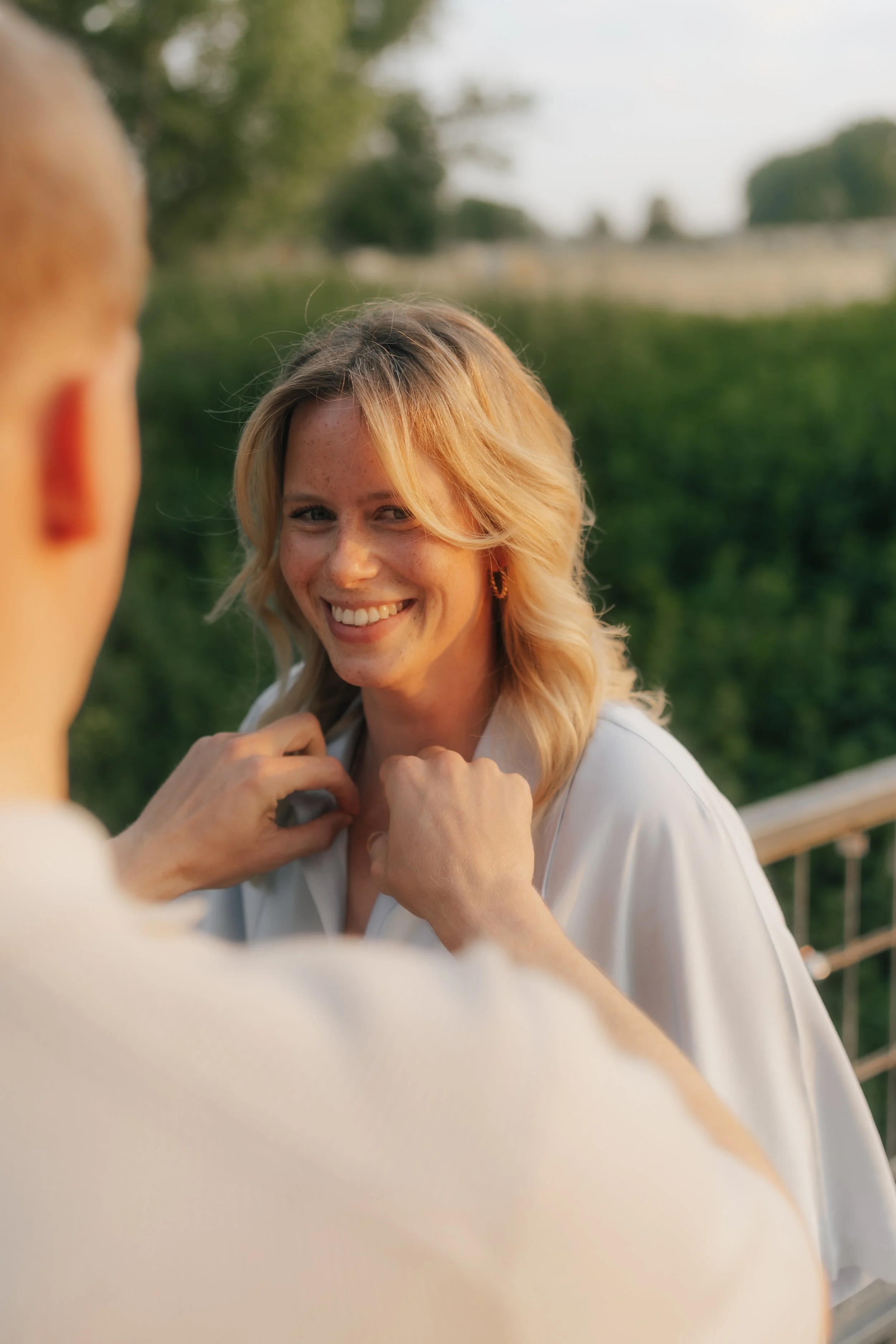 Eine lachende blonde Frau im weißen Hemd, die sich auf einer Brücke befindet, im Sonnenlicht bei Sonnenuntergang, umgeben von grünen Bäumen.