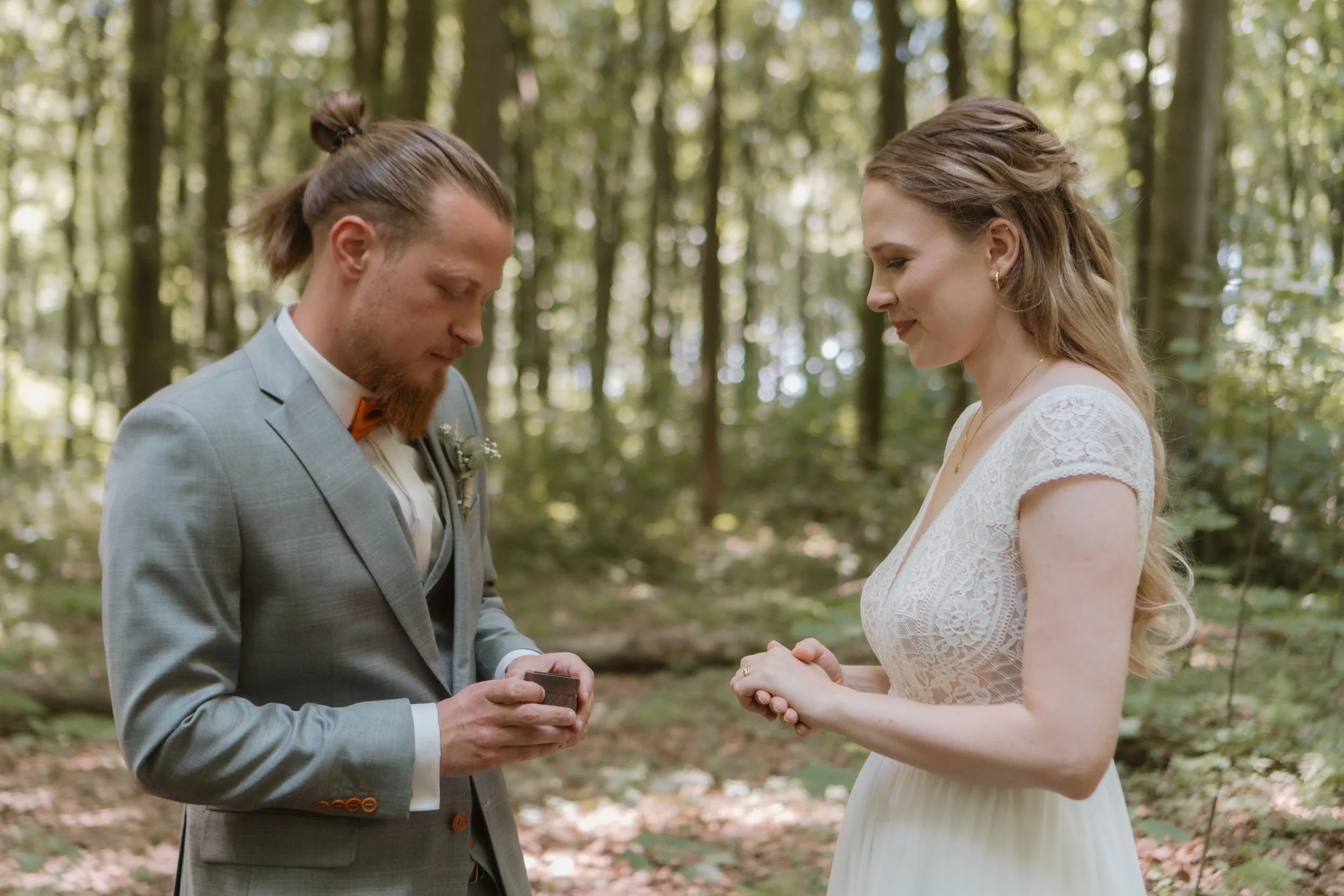 Ein Brautpaar bei einer Zeremonie im Wald, der Bräutigam hält einen Ring, die Braut sieht erleichtert und glücklich aus.
