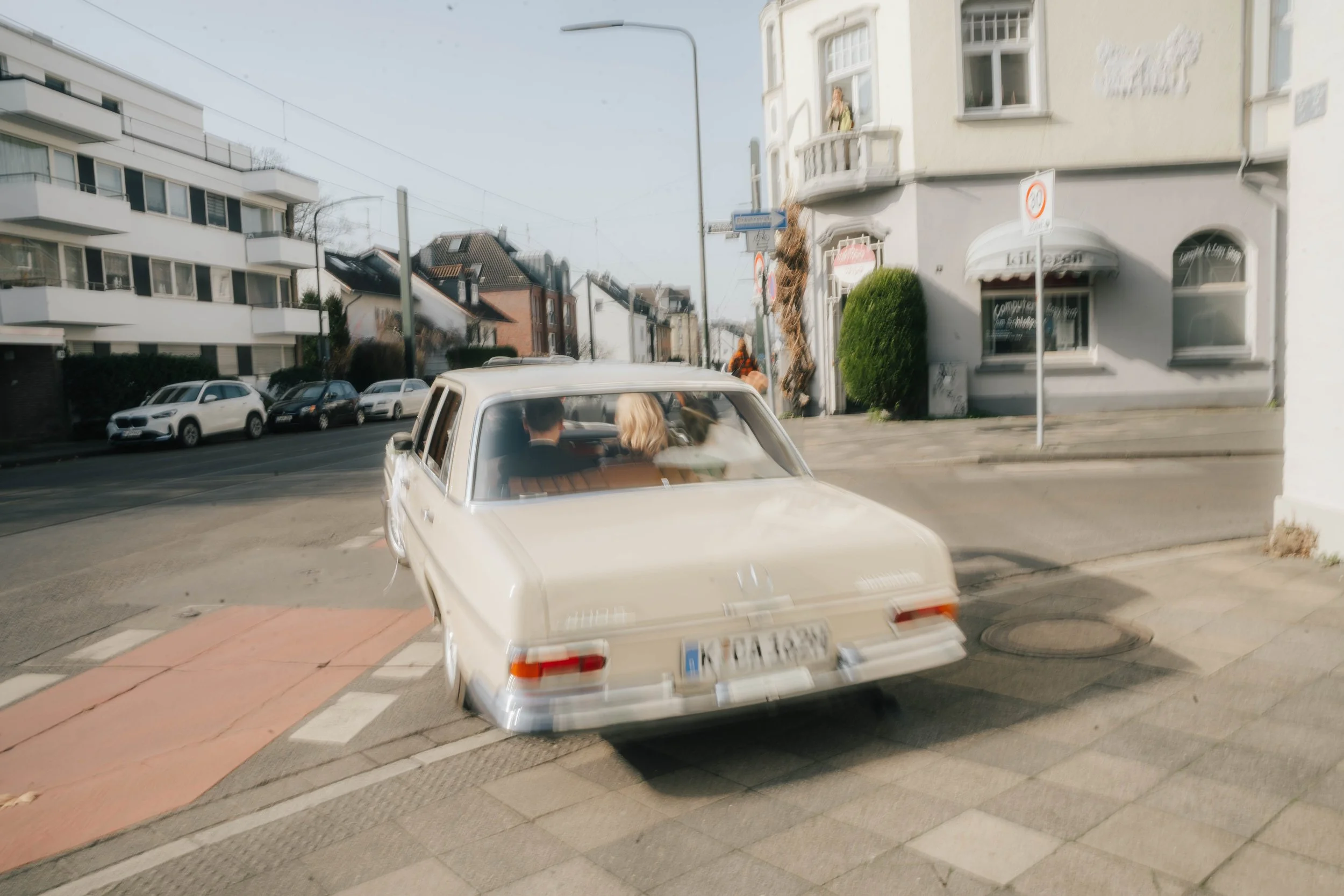Ein weißes Auto fährt an einer Kreuzung in einer städtischen Gegend vorbei. Im Hintergrund sind mehrstöckige Gebäude, parkende Autos und Fußgänger zu sehen.