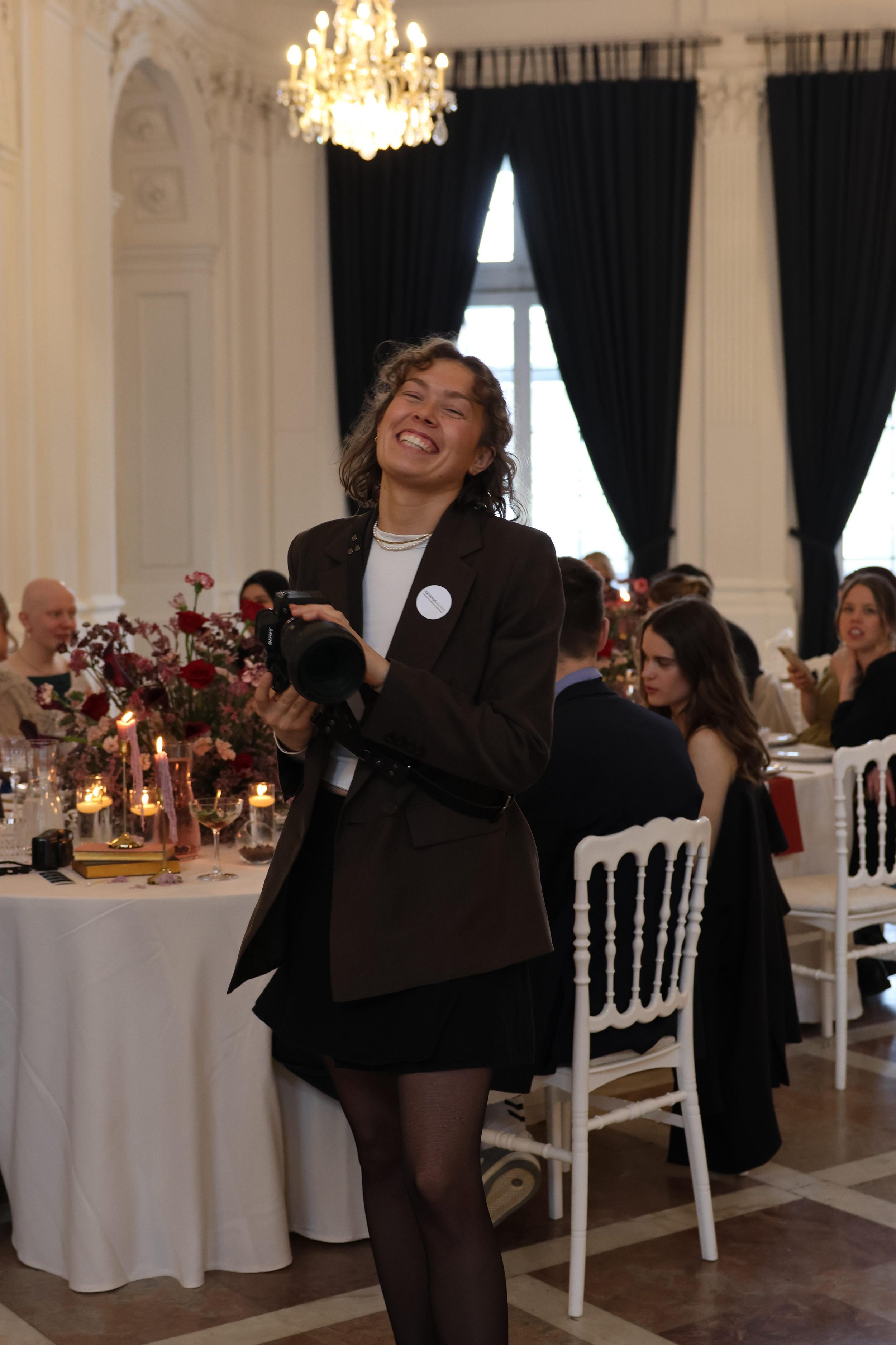 Junge Frau lacht bei einem festlichen Abendessen in einem eleganten Raum mit schwarzen Vorhängen, Kristallleuchter und Tafel mit Kerzen und Blumen.
