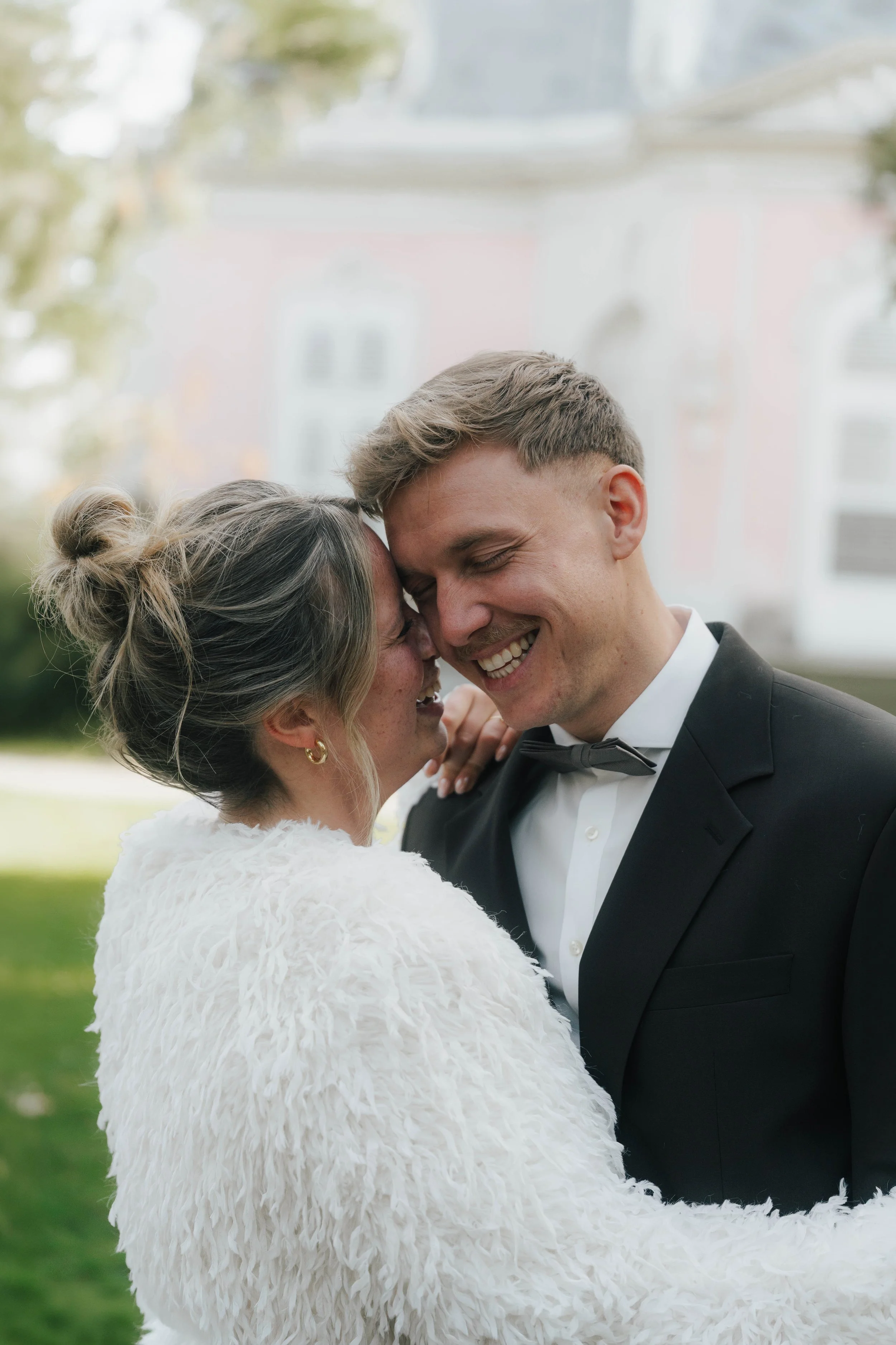 Ein lachendes Paar bei einer Hochzeit, der Mann trägt einen schwarzen Anzug und eine Fliege, die Frau trägt ein weißes Hochzeitskleid mit Fell-Details, sie sind im Freien.