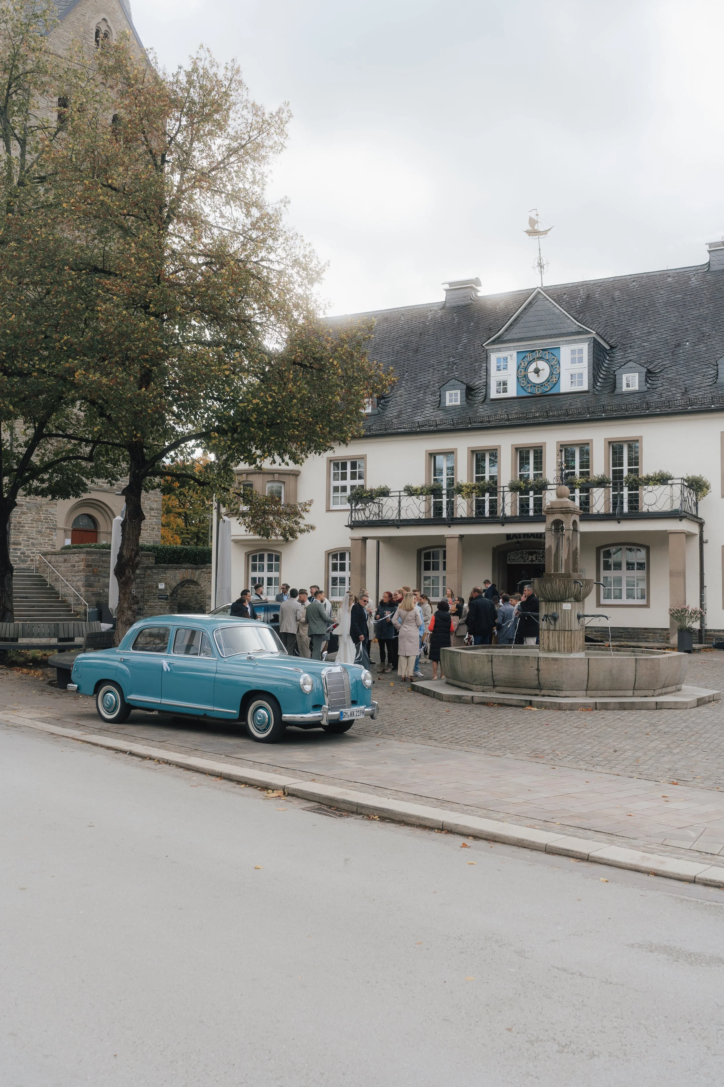 Eine Kutsch mit klassischen Design vor einem Platz mit Menschen, Brunnen und einem alten Gebäude mit Uhrturm und Wetterfahne.