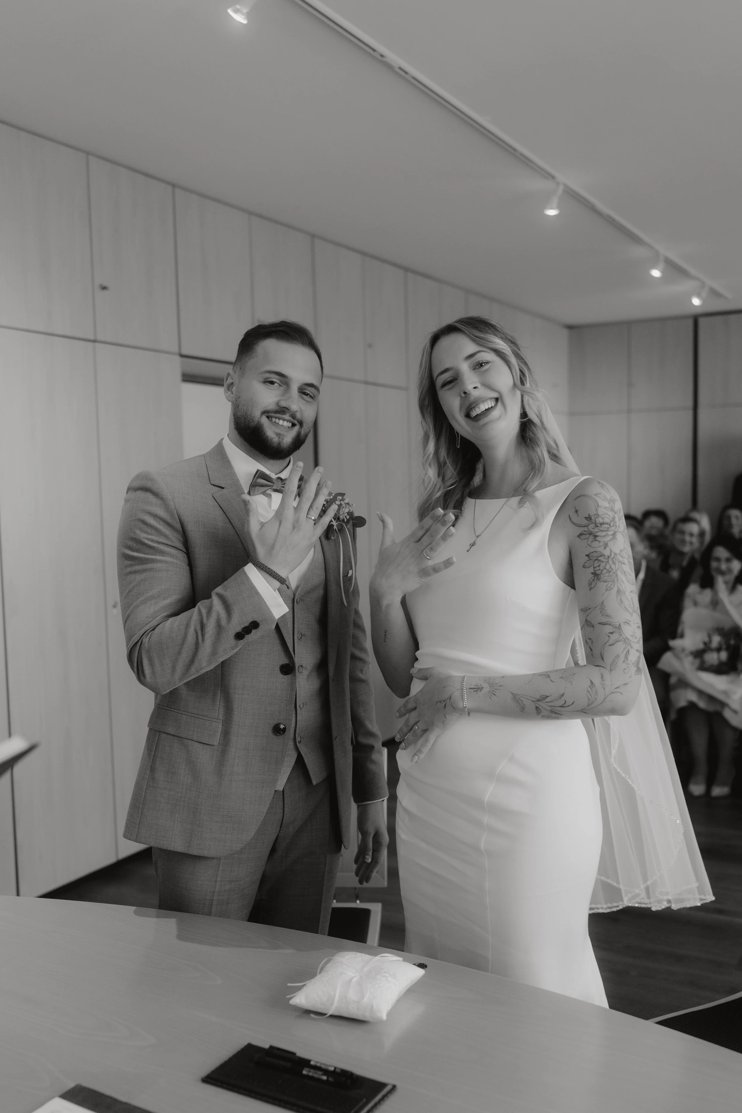 Ein Brautpaar zeigt ihre Eheringe bei einer Trauung in einem Raum mit Holzpaneelen, umgeben von Gästen.