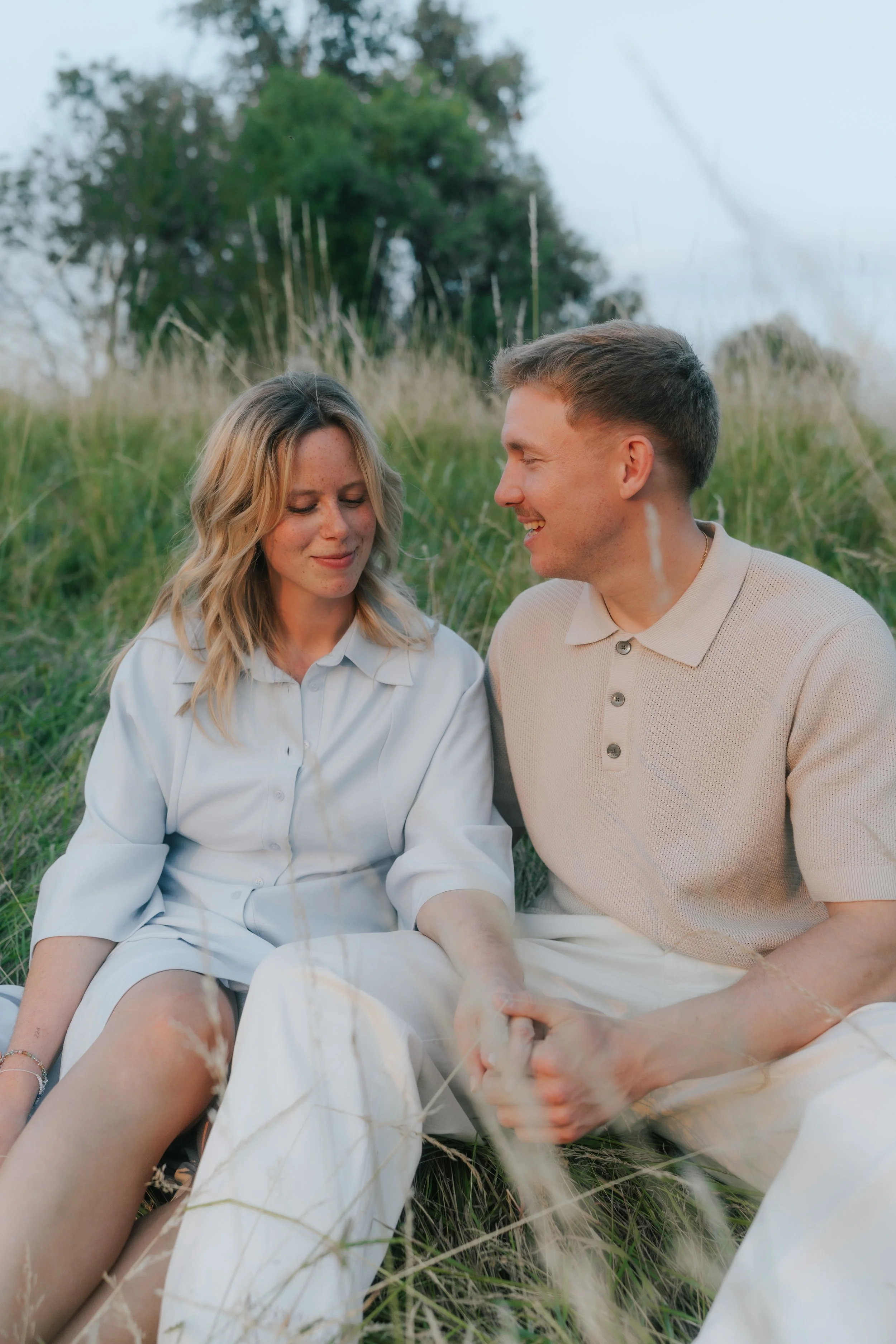Ein Paar sitzt zusammen im Gras in einer Naturumgebung, hält Händchen und lächelt sich liebevoll an.
