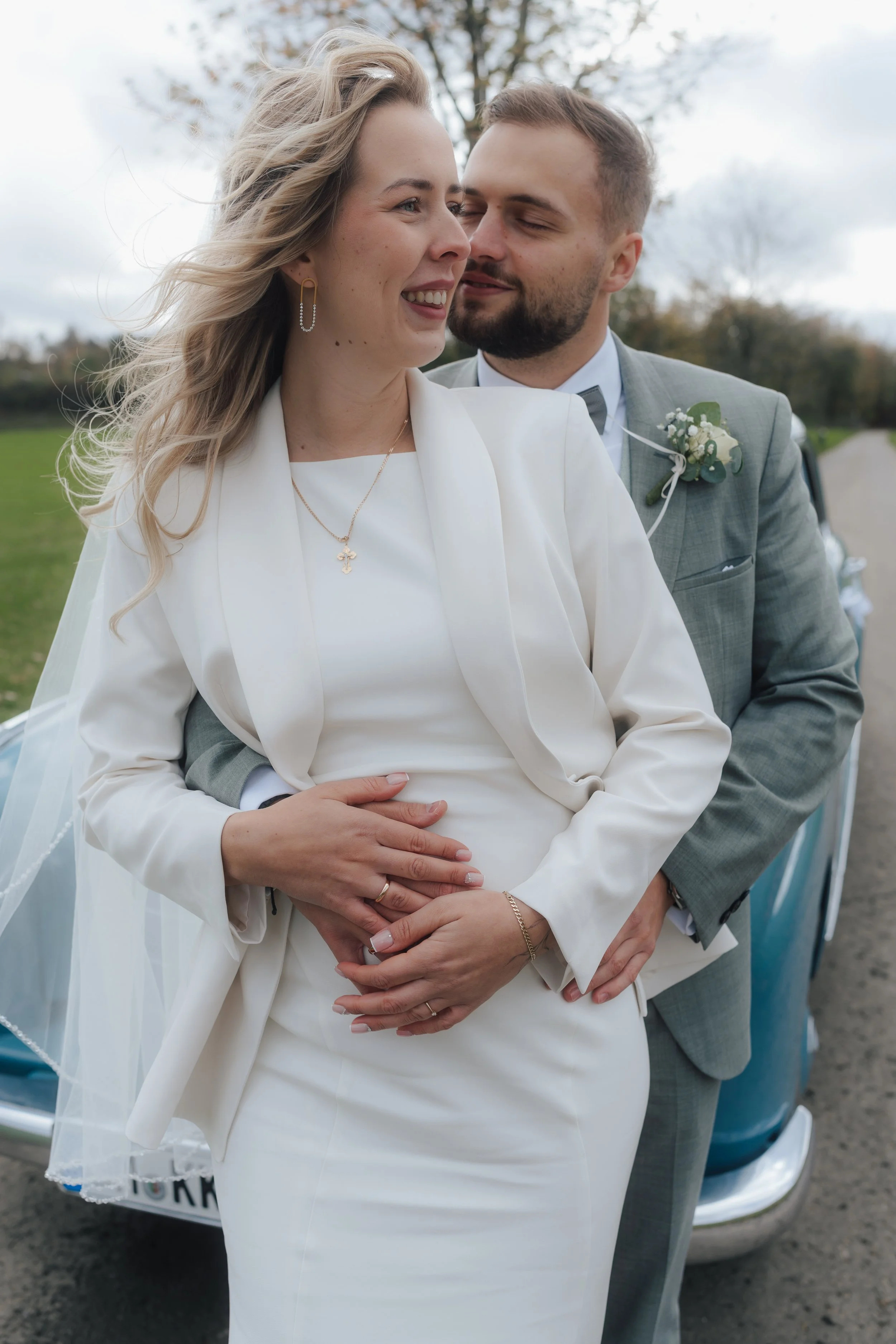 Ein glückliches Brautpaar umarmt sich vor einem Oldtimer, im Freien bei bewölktem Himmel, während sie an ihrer Hochzeit teilnehmen.