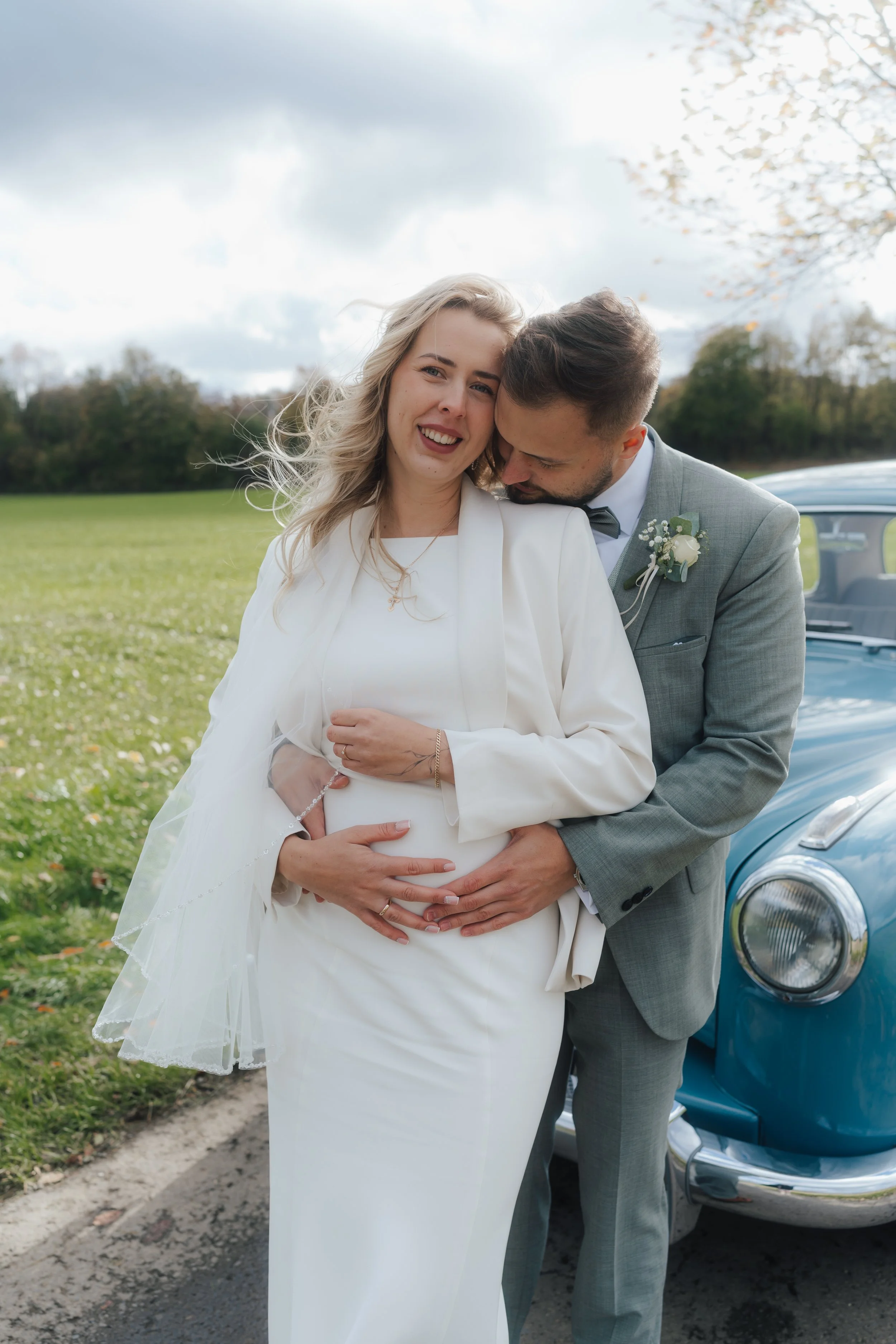 Ein glückliches frisch verheiratetes Paar bei einem Fotoshooting im Freien, Frauen in weißem Kleid, Männer im grauen Anzug, vor einem blauen Oldtimer, auf einer Wiese mit Bäumen bei bewölktem Himmel.