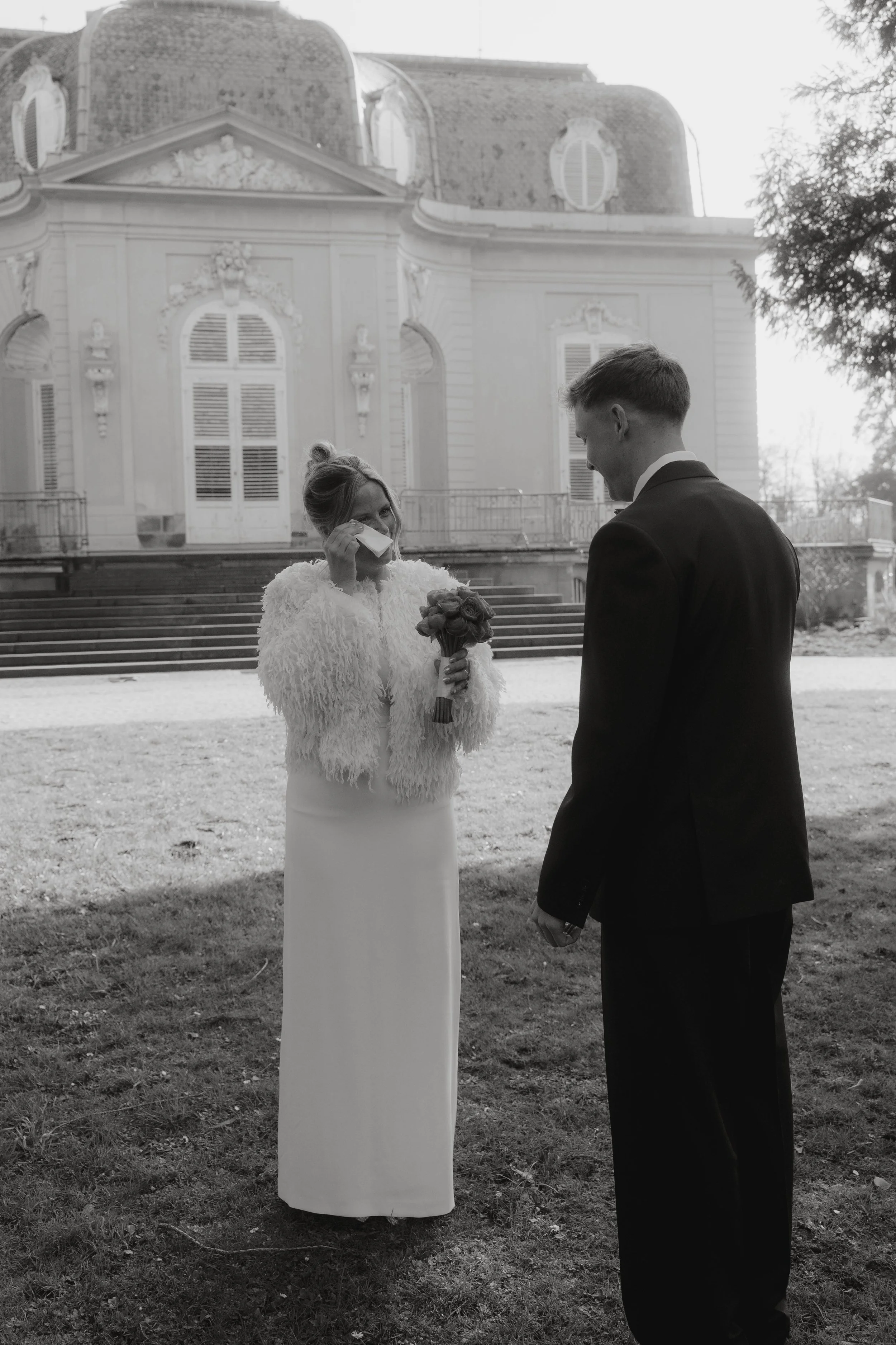 Ein Hochzeitsfoto von einer Frau mit Brautkleid und einer Man mit Anzug, die sich vor einem alten Gebäude im Freien befinden, während die Frau weint und die Frau dem Mann einen Ring an den Finger steckt.