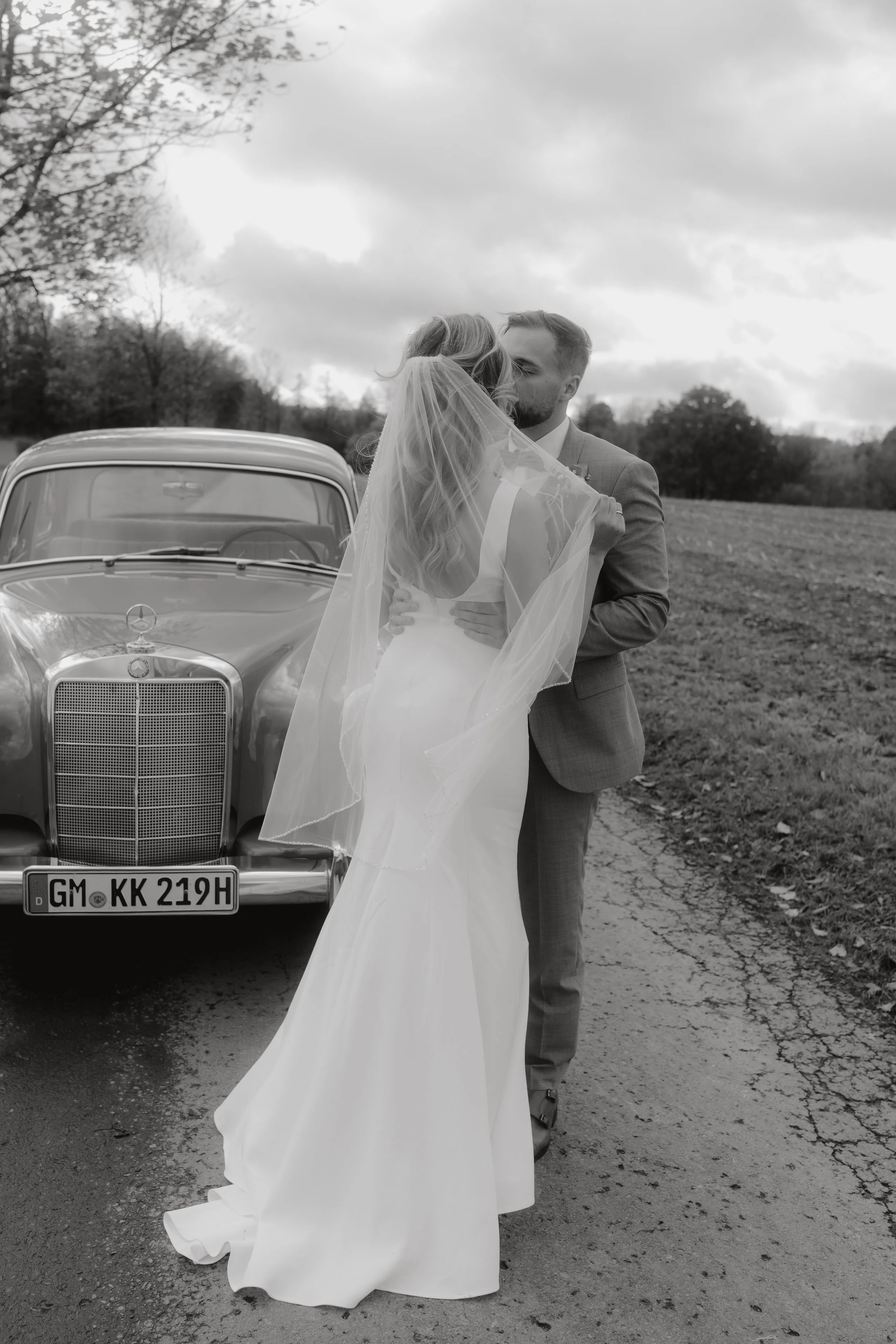 Braut und Bräutigam küssen sich vor einem alten Mercedes-Benz auf einer Landstraße, im Hintergrund Bäume und Wolken am Himmel, Schwarz-Weiß-Fotografie.