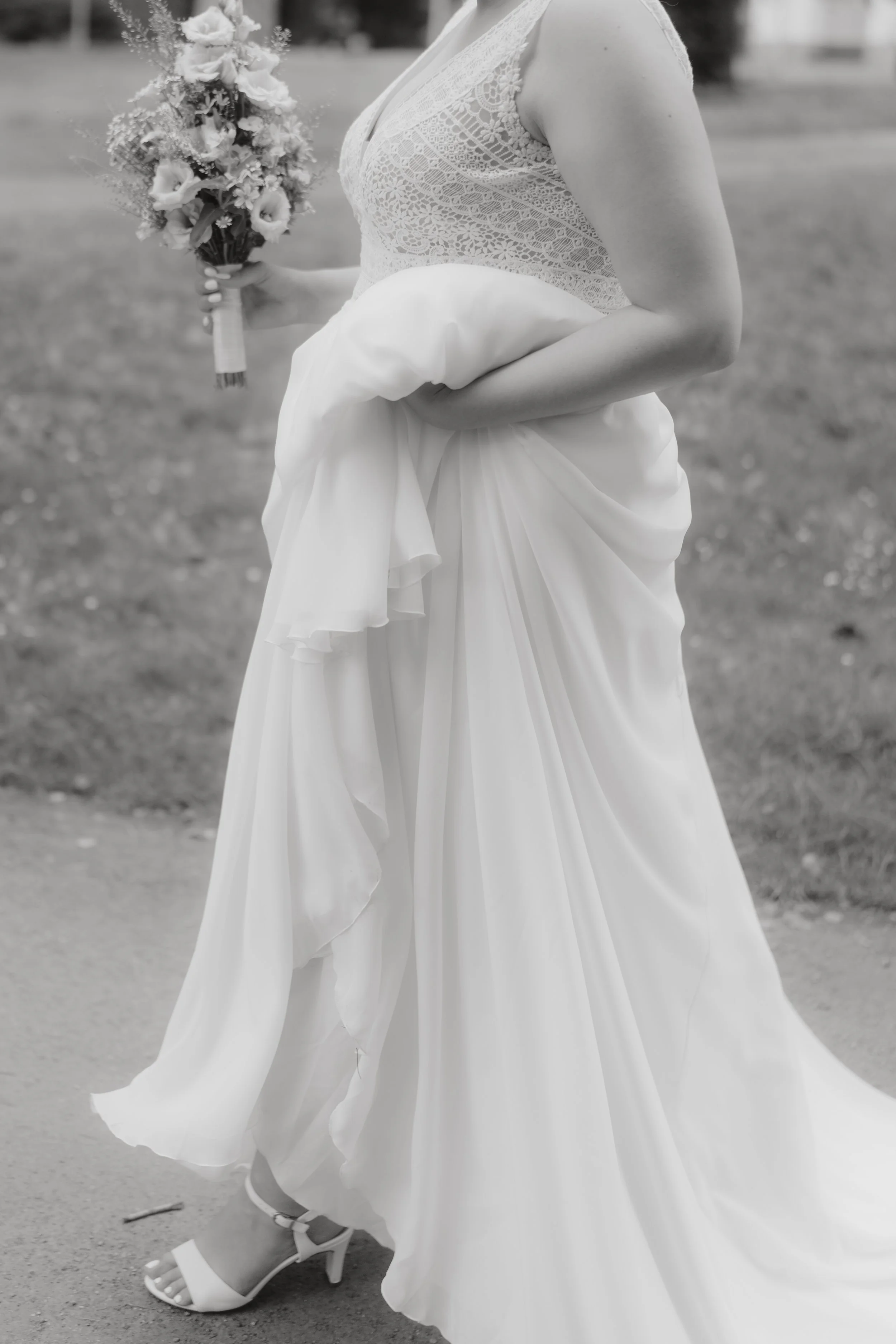 Eine Braut in einem weißen Hochzeitskleid hält einen Blumenstrauß in der linken Hand, die rechte Hand hebt den unteren Teil ihres Kleides. Das Bild ist in Schwarz-Weiß.