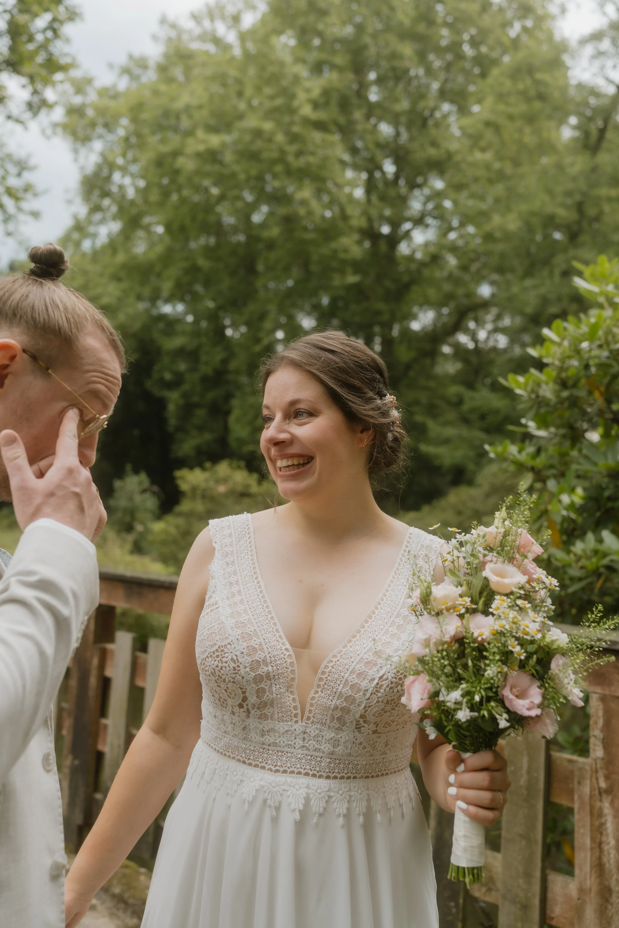 Eine lachende Braut mit Brautstrauß, im Gespräch mit einem Mann, an einem grünen, bewaldeten Ort.