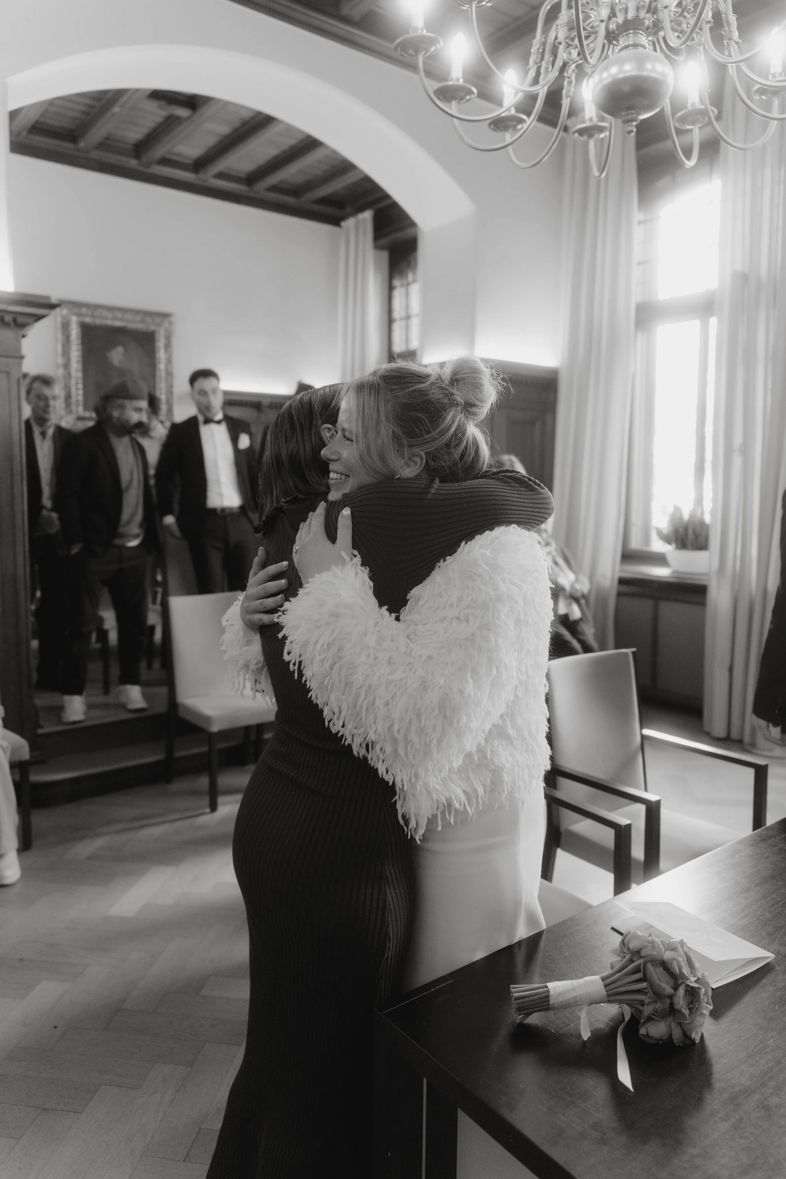 Zwei Frauen umarmen sich herzlich in einem festlich dekorierten Raum, im Hintergrund stehen mehrere Männer in formeller Kleidung. Es handelt sich vermutlich um eine Feier oder Zeremonie.