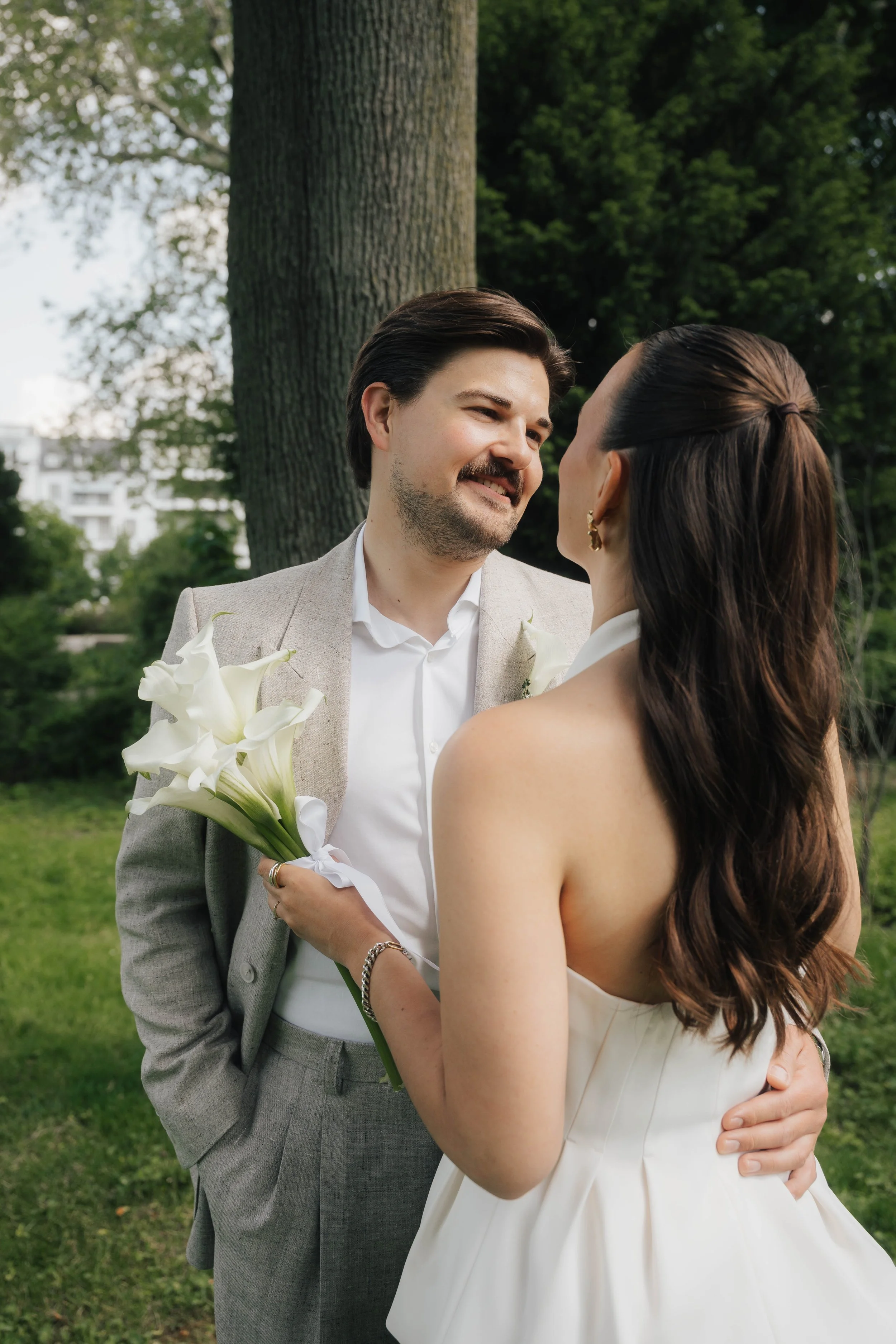 Ein Mann und eine Frau stehen sich bei einer Hochzeit im Freien gegenüber, der Mann hält einen Blumenstrauß und beide lächeln sich an.