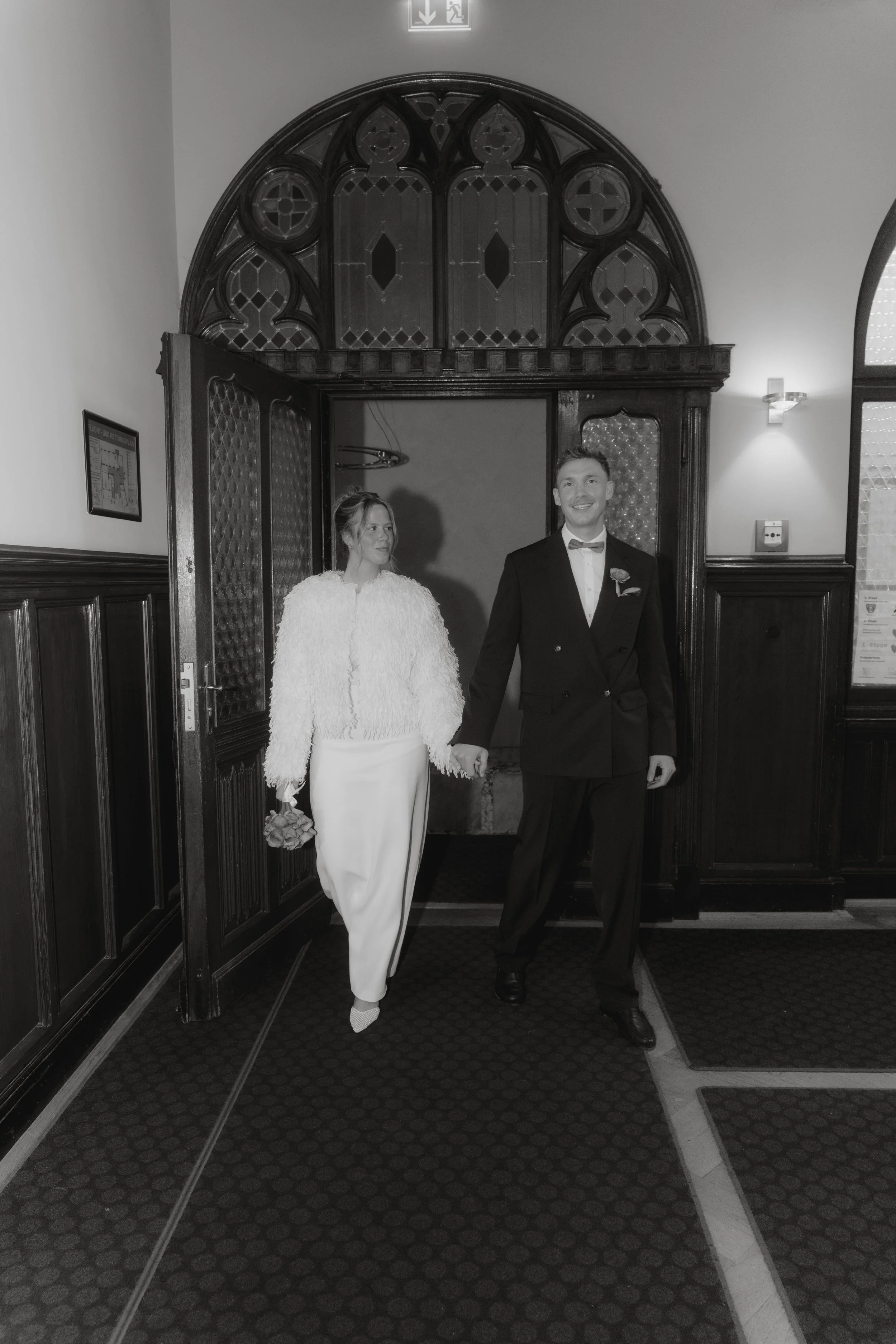 Ein Mann in Smoking und eine Frau in einem weißen Kleid halten die Hände und gehen durch eine Tür in einem historischen Gebäude, vermutlich bei einer Hochzeitszeremonie.
