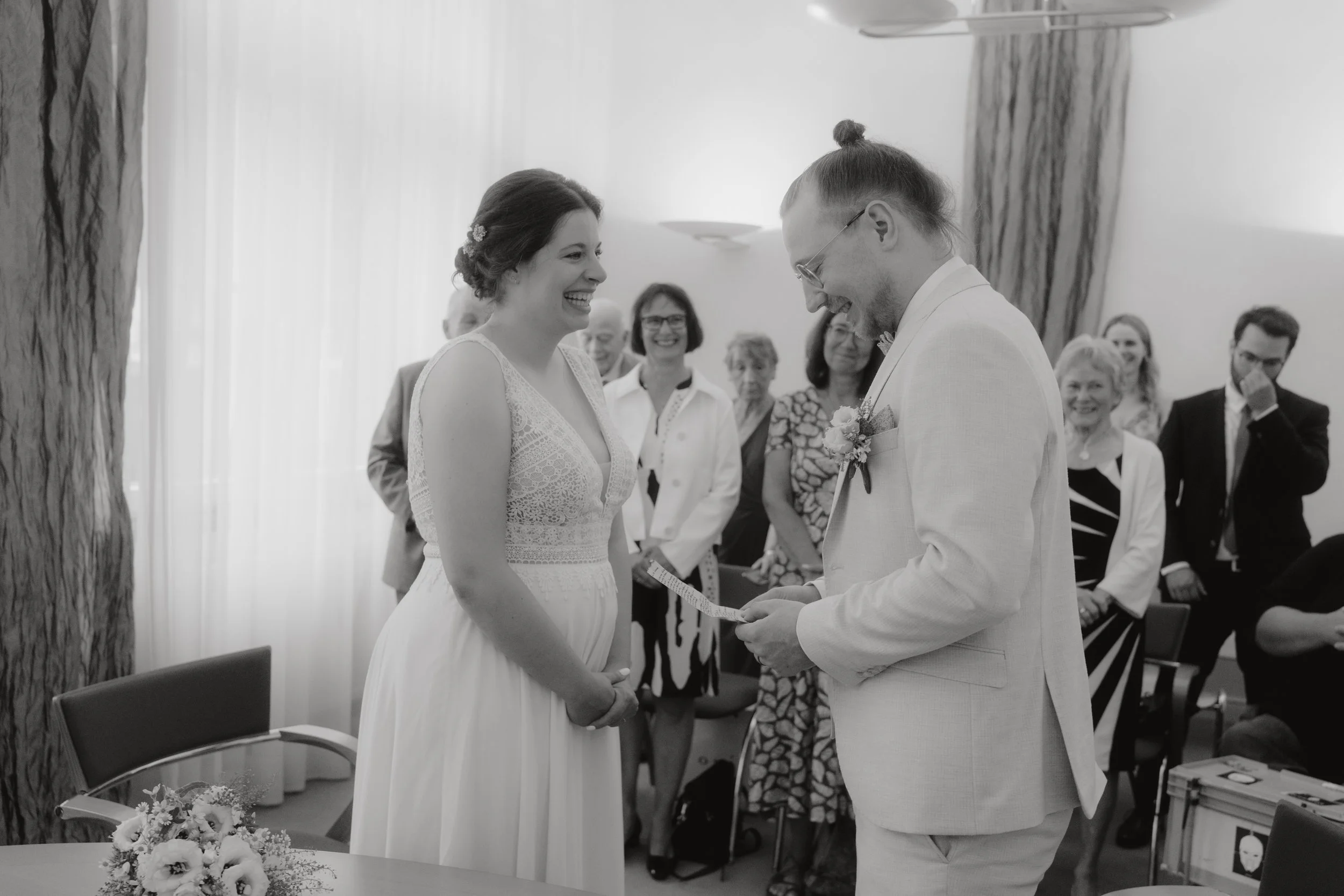 Ein Brautpaar bei ihrer standesamtlichen Trauung, lachend und sich gegenüberstehend, während der Ehemann dem/der/die Hochzeitsstandesbeamte/in eine Urkunde übergibt, mit Gästen im Hintergrund.