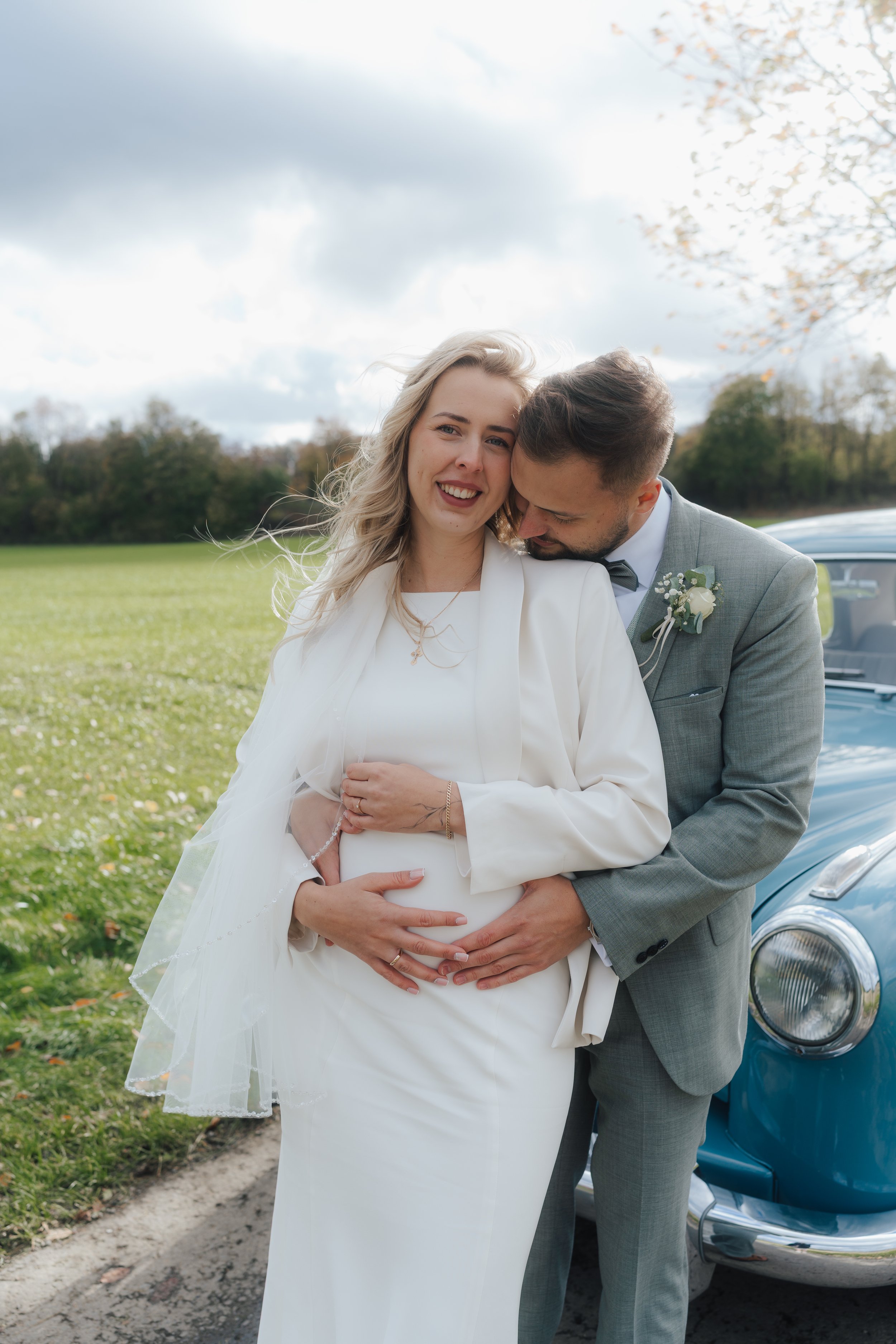 Ein glückliches Hochzeitspaar, ein Mann und eine schwangere Frau, posieren vor einem klassischen blauen Auto auf einer grünen Wiese mit Bäumen, bei bewölktem Himmel.