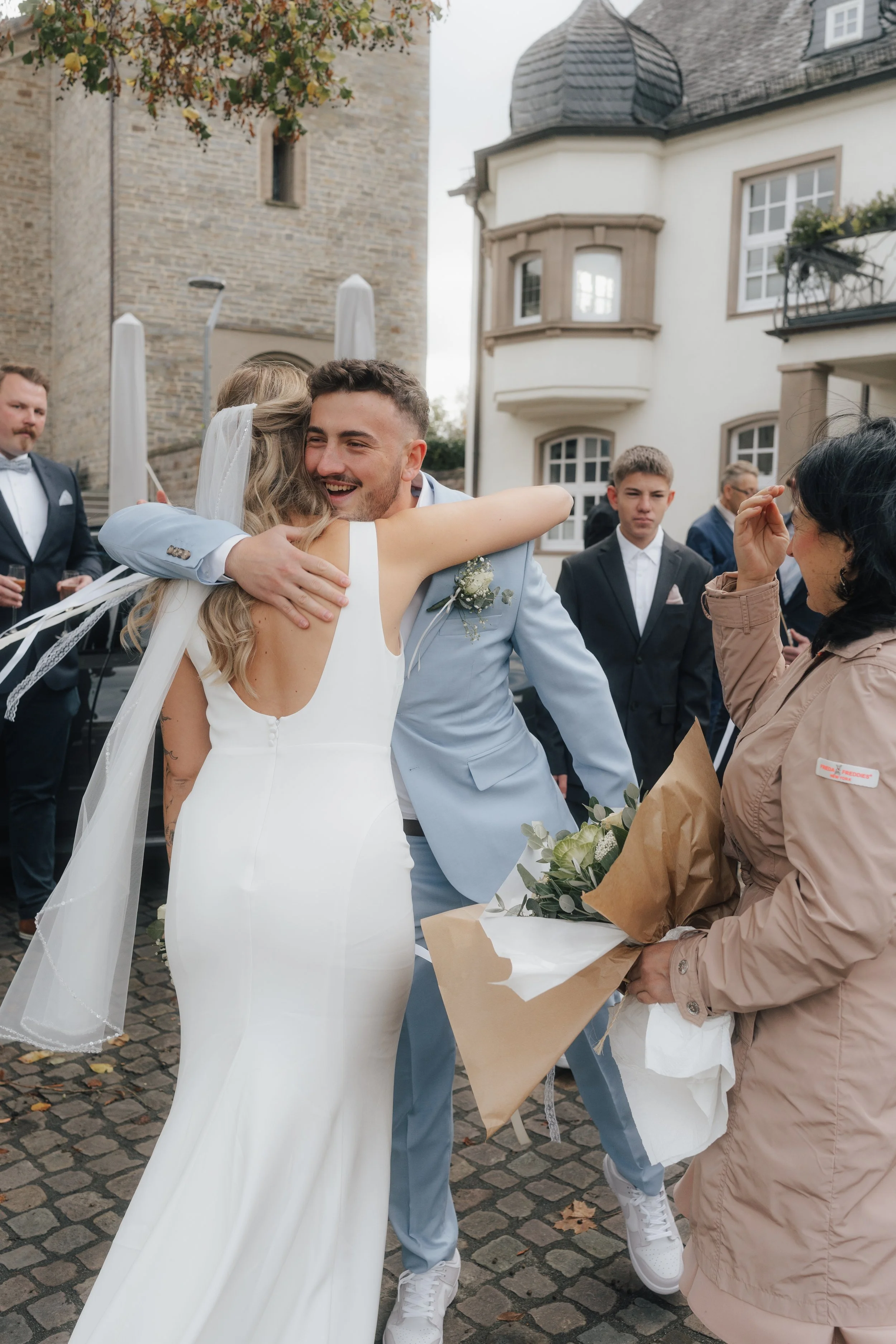 Ein Brautpaar umarmt sich bei einer Hochzeit im Freien, umgeben von Gästen, einige im Anzug, mit historischen Gebäuden im Hintergrund.