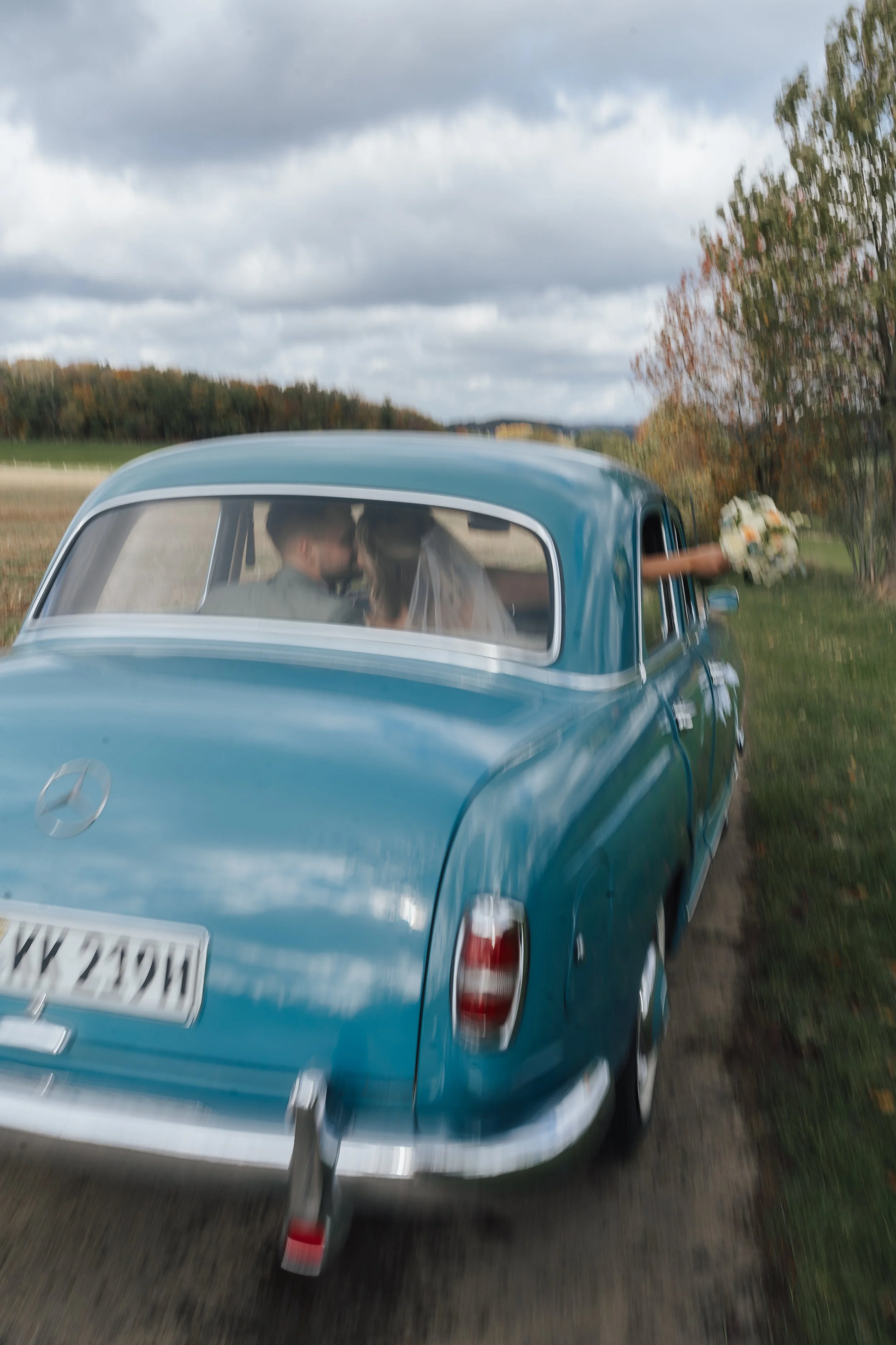 Ein blauer Oldtimer, in den ein Paar sitzt, während eine Person mit Blumen herausholt, auf einer ländlichen Straße mit bewölktem Himmel im Hintergrund.