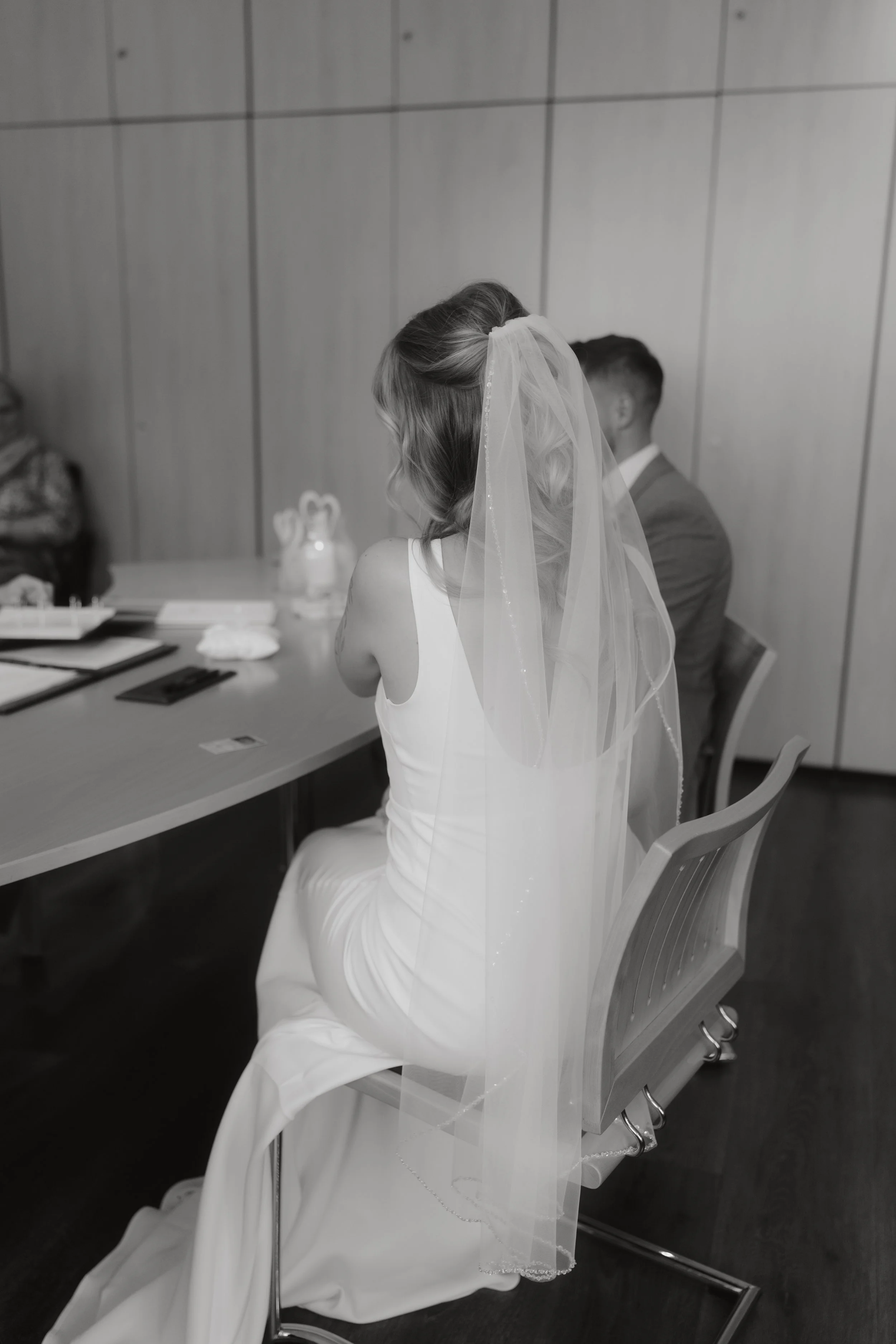 Schwarz-weiß-Foto einer Braut in einem weißen Kleid mit Schleier, die auf einem Stuhl sitzt, im Hintergrund Mann in Anzug in einem Raum mit Holzverkleidung an den Wänden