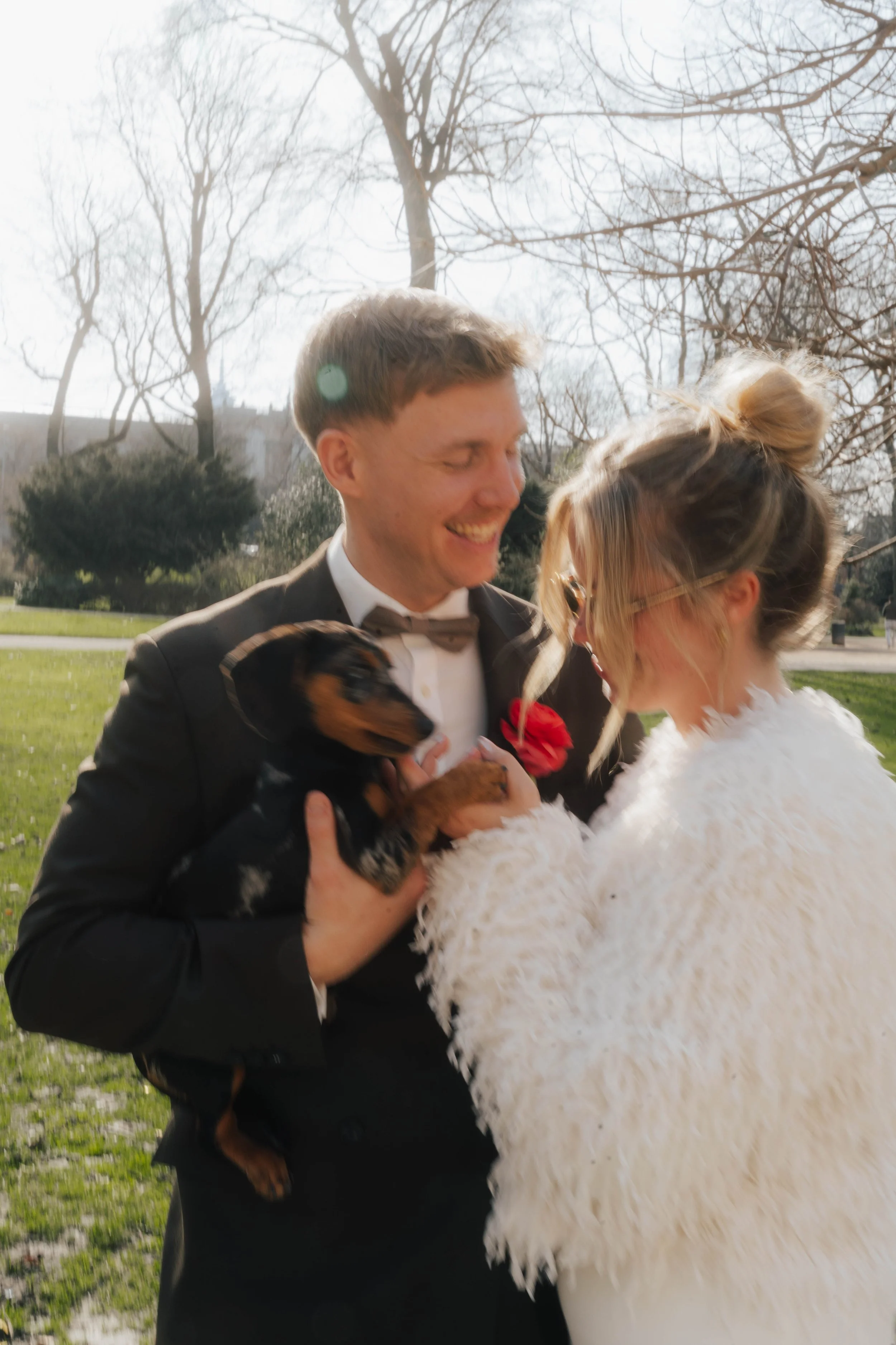 Ein Mann und eine Frau, die sich auf einer Wiese im Freien nahe Bäume befinden, lachen und halten ein Hundewelpen in den Armen.