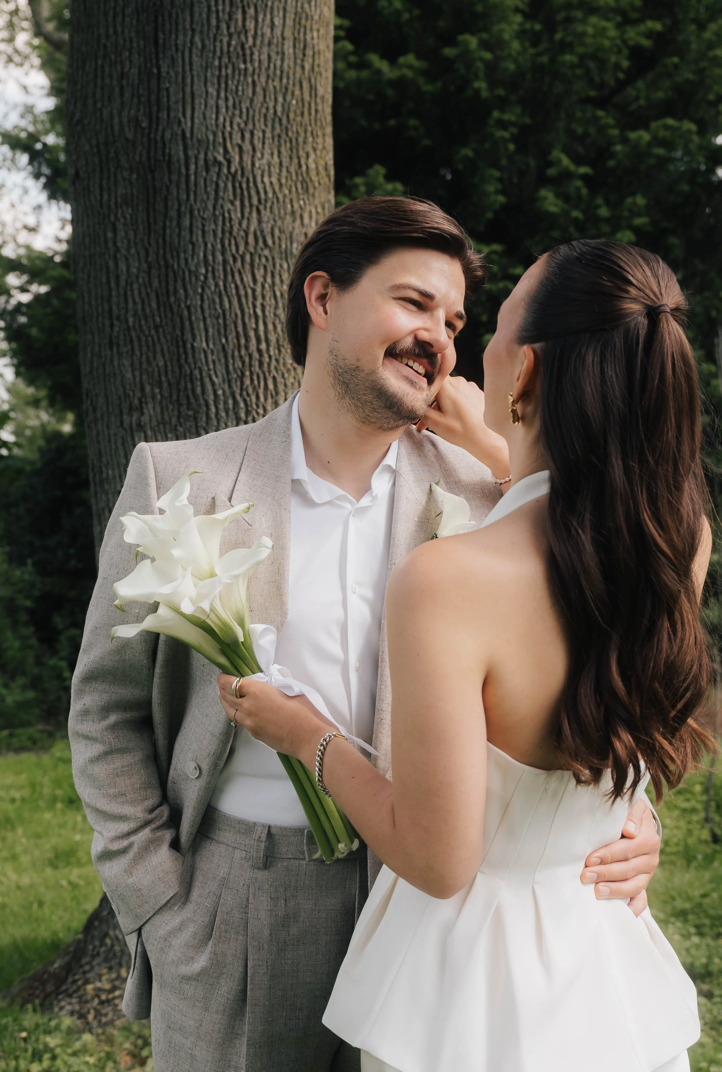 Ein glückliches Brautpaar steht im Park, umarmt sich und lächelt einander an, während die Braut eine weiße Hochzeitsskleid trägt und den Bräutigam mit einem Anzug. Sie hält einen Blumenstrauß aus Calla-Lilien. Im Hintergrund sind Bäume und grüne Natu