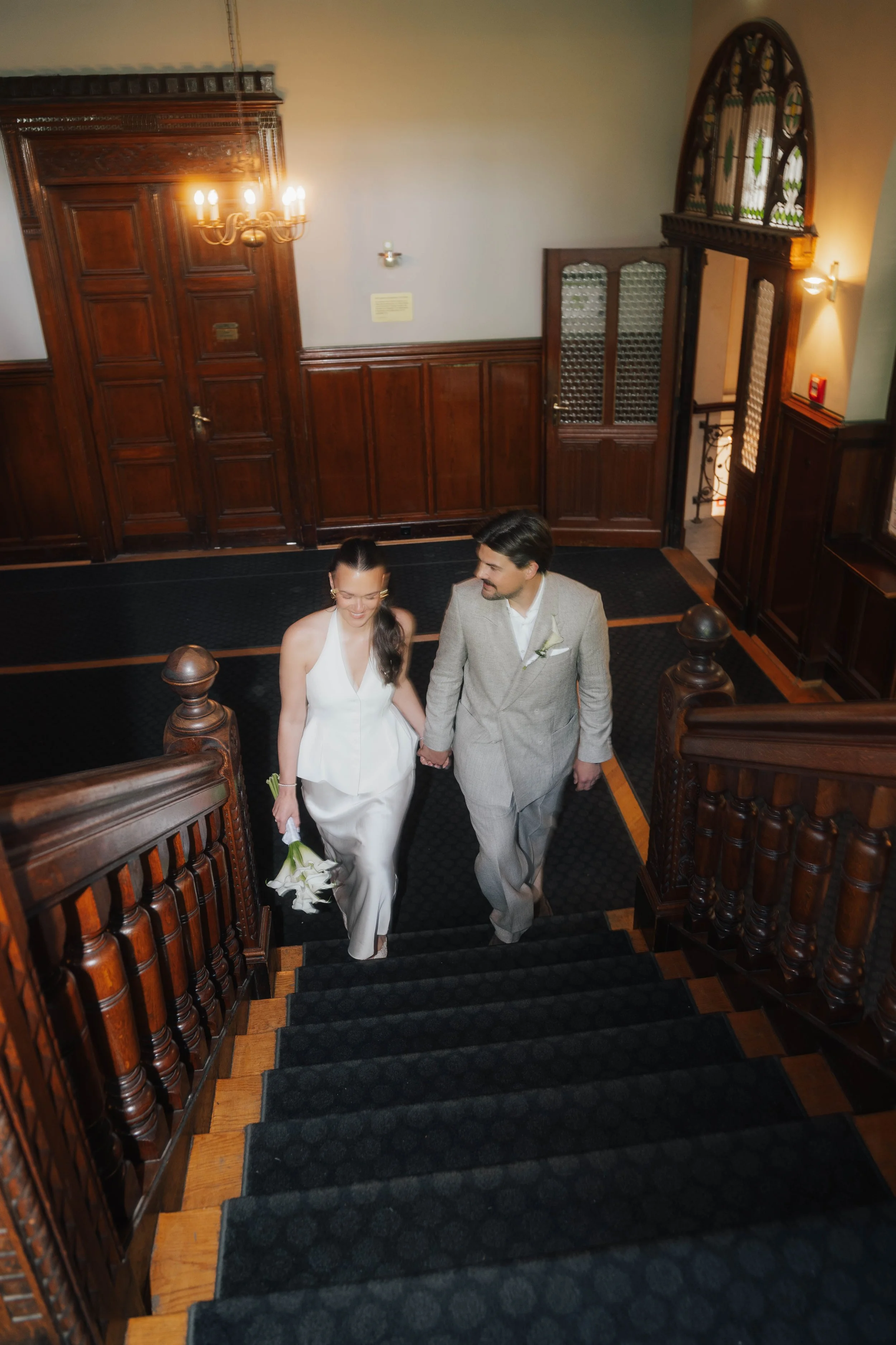 Ein Paar, ein Mann in einem Hellgrauen Anzug und eine Frau in einem weißen Kleid, geht die Treppe eines historischen Gebäudes hinauf. Sie lachen und halten sich an den Händen. DieTreppengeländer sind aus dunklem Holz, die Wände sind ebenfalls mit Hol
