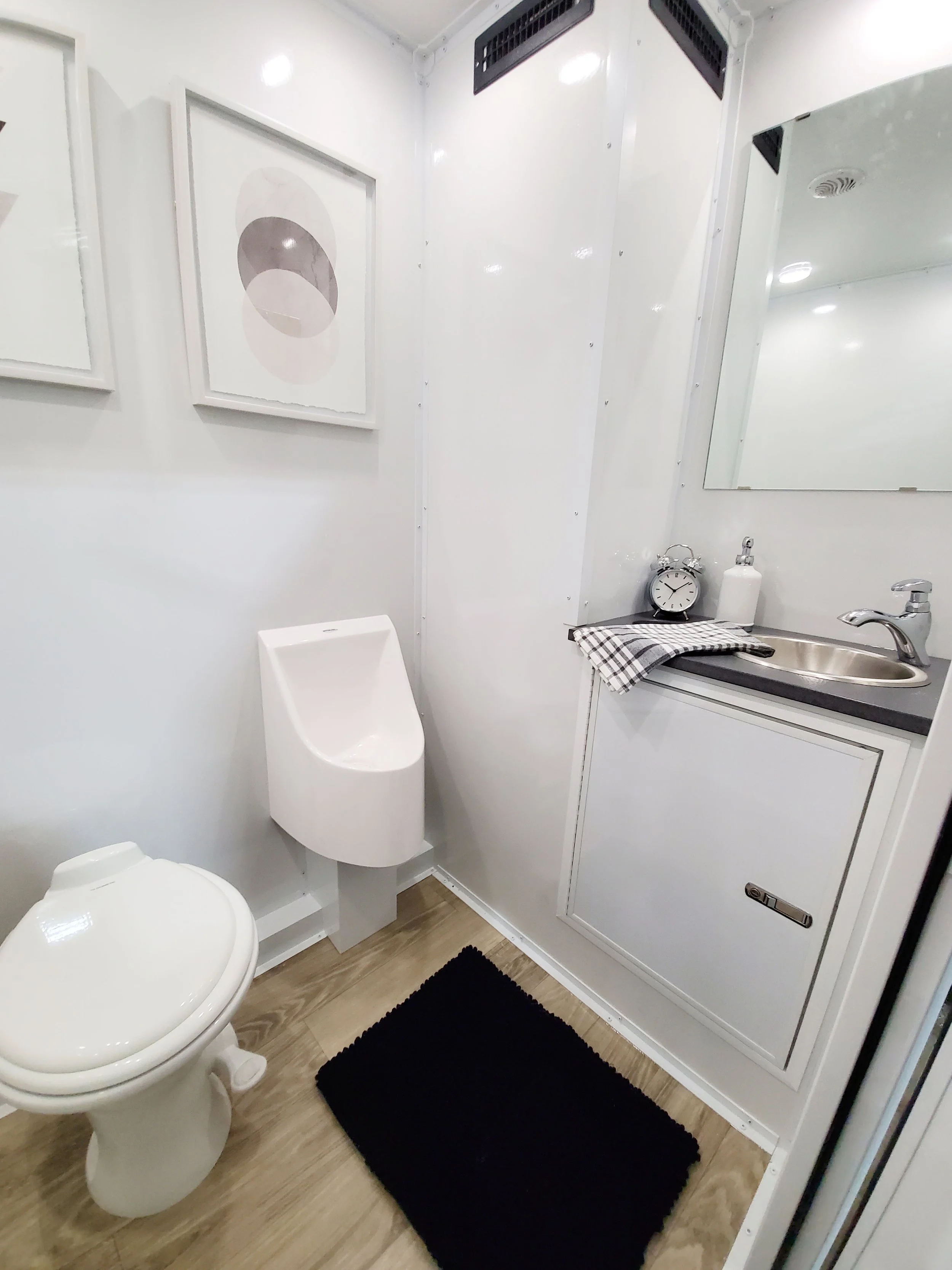 A modern bathroom inside a restroom trailer featuring a white toilet, a built-in urinal, a small sink with a mirror above, black and white decor, framed abstract art, and a dark rug on a wooden floor.
