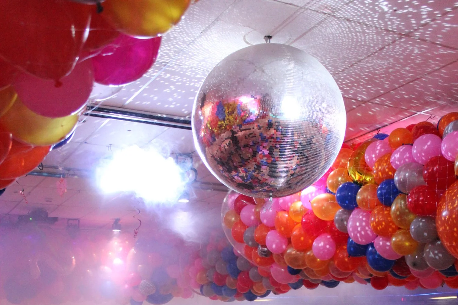 A disco ball hanging from the ceiling reflecting colorful lights, with clusters of pink, orange, yellow, blue, and gray balloons surrounding it, and a bright light illuminating the scene.